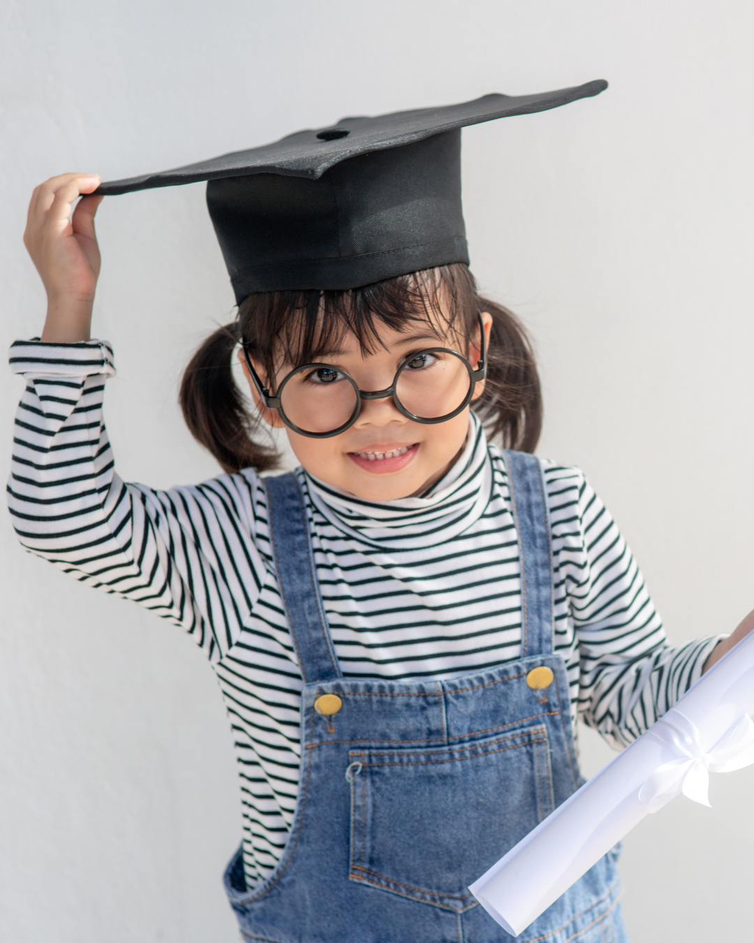 A young girl wearing glasses, a striped long-sleeve shirt, and denim overalls, holding a rolled-up document in one hand, and adjusting a graduation cap on her head with the other, standing against a plain wall.