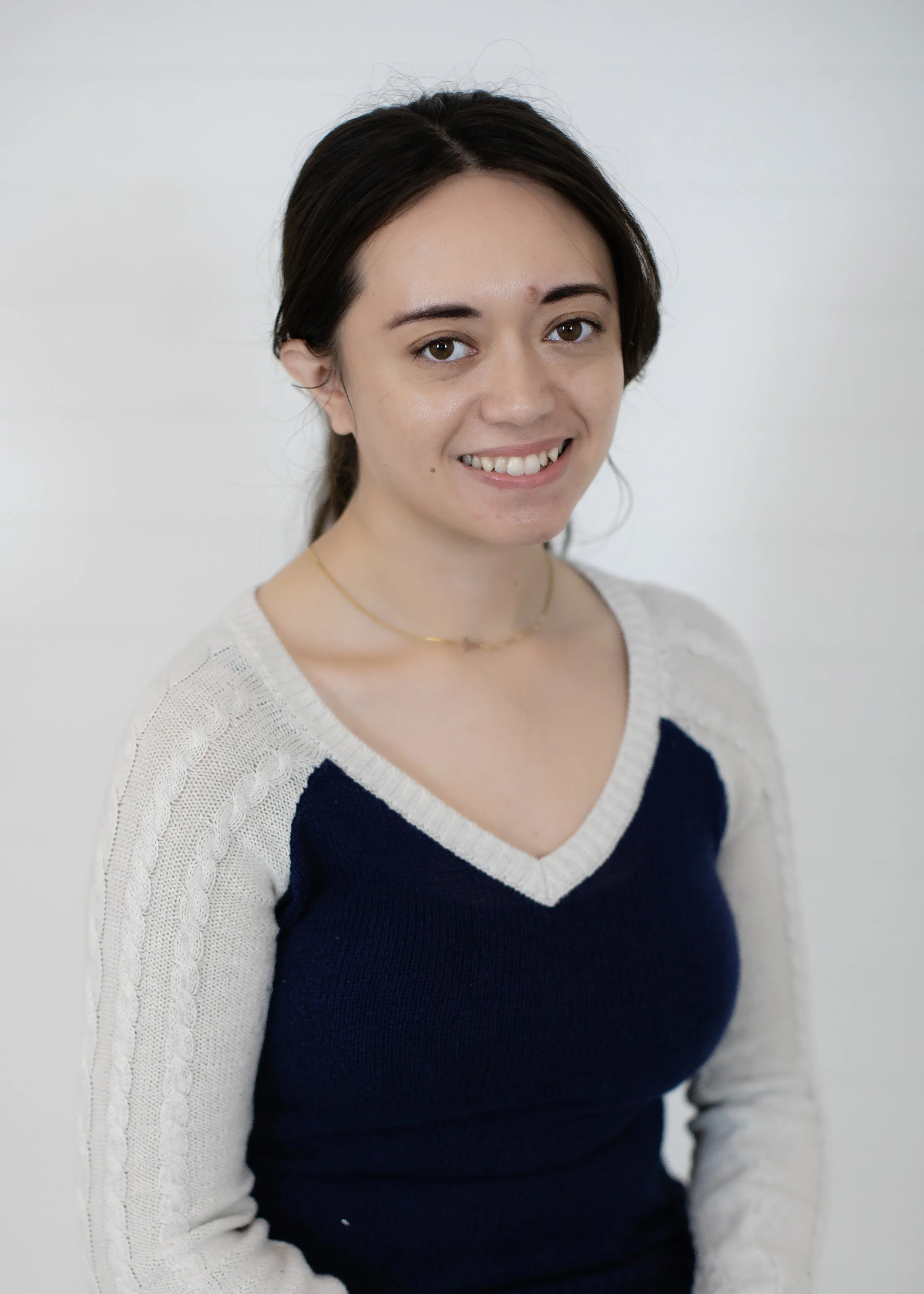 Portrait of a young woman with dark brown hair, wearing a cream and navy blue sweater, and a small gold necklace, smiling against a plain background.