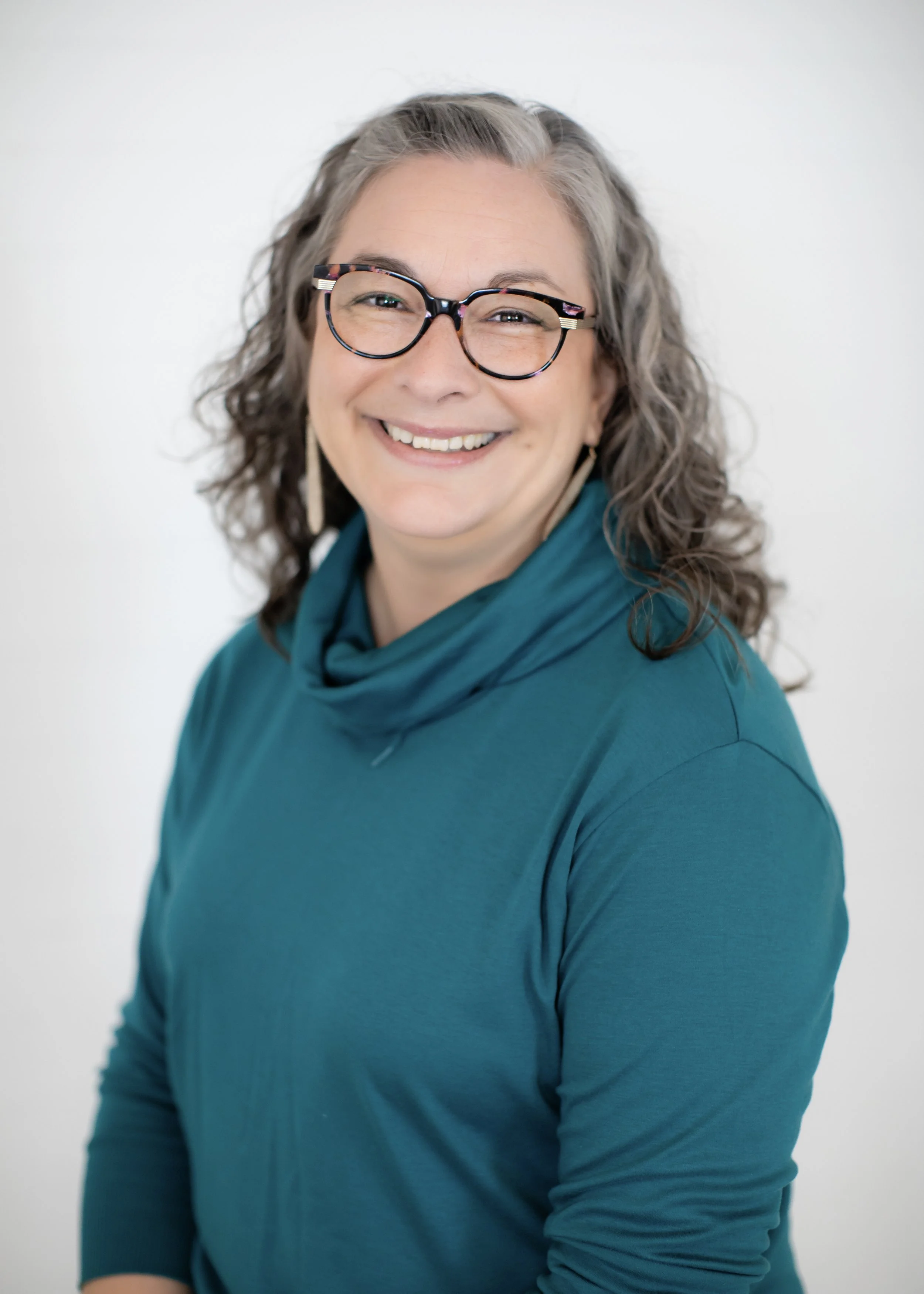 A woman with curly, gray hair and glasses smiling in front of a plain white background, wearing a teal long-sleeve top.