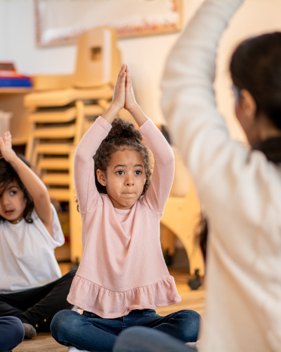 A young girl with curly hair and a pink long-sleeve shirt sits cross-legged on the floor in a classroom while raising her hands above her head in a Yogi pose. Other children are nearby, also participating, and a teacher or instructor is leading the activity.
