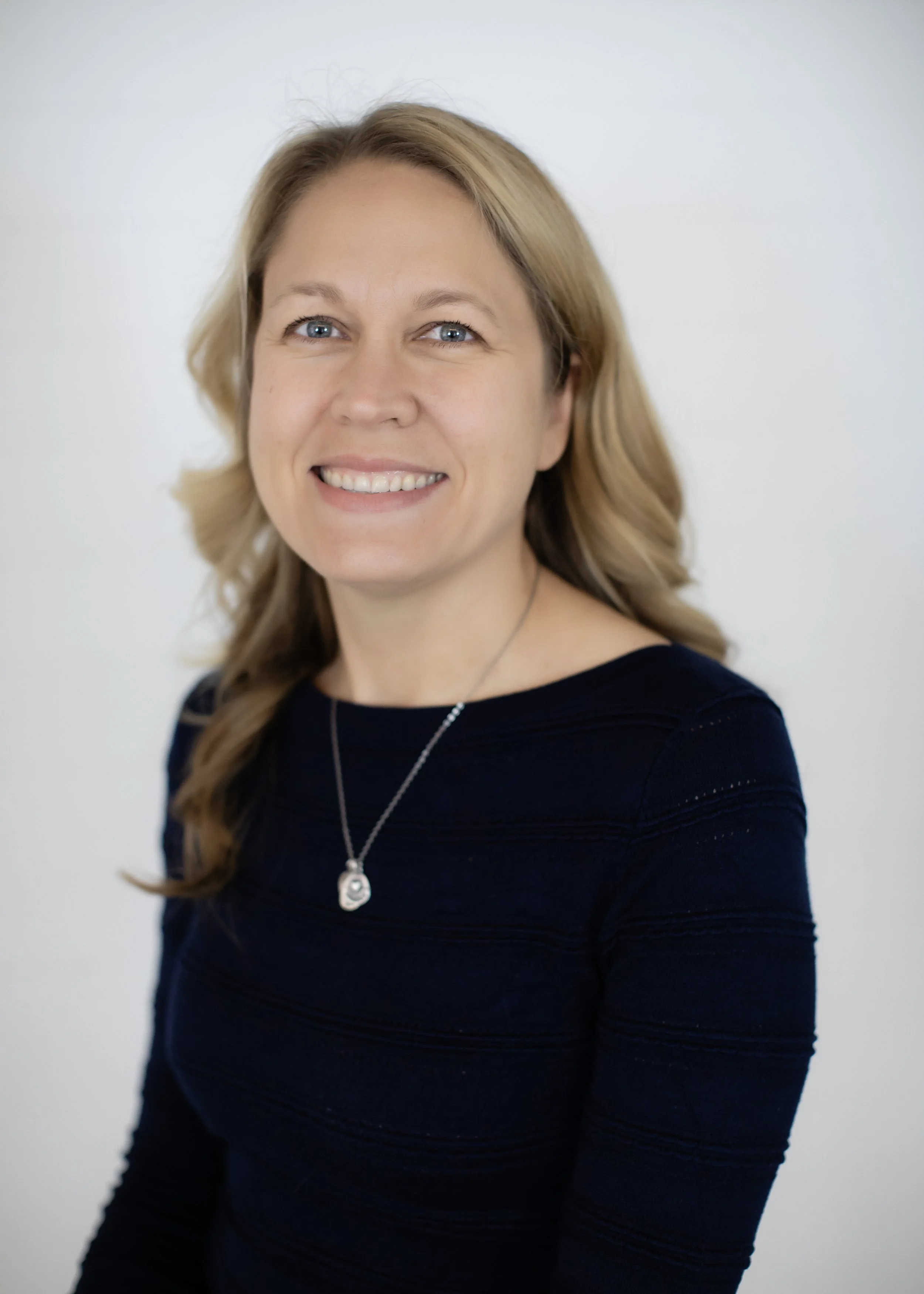 A woman with blonde hair, blue eyes, and a big smile is wearing a black top and a silver necklace. She is standing against a plain white background.