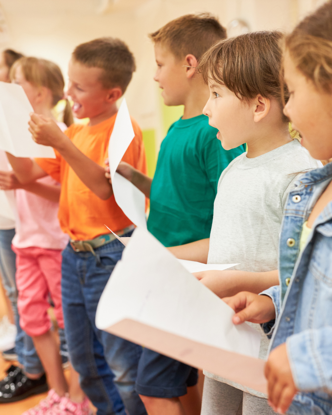 Children standing in a row, holding papers, singing or speaking, indoors.