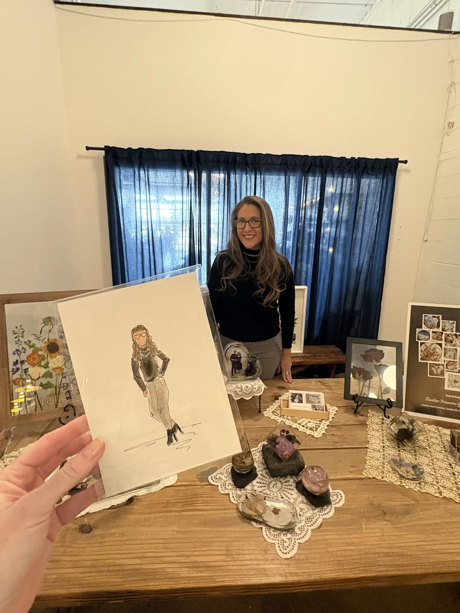 A woman with long hair and glasses smiling at a table with various rocks and art pieces, with a hand holding a illustrated card in the foreground.