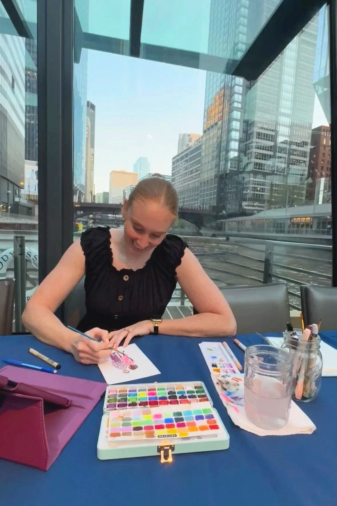 A woman sitting at a table in a modern glass-walled building, painting a picture of a woman in a pink dress. The table has watercolor supplies, markers, and a glass of water. The city skyline with tall buildings is visible through the windows.