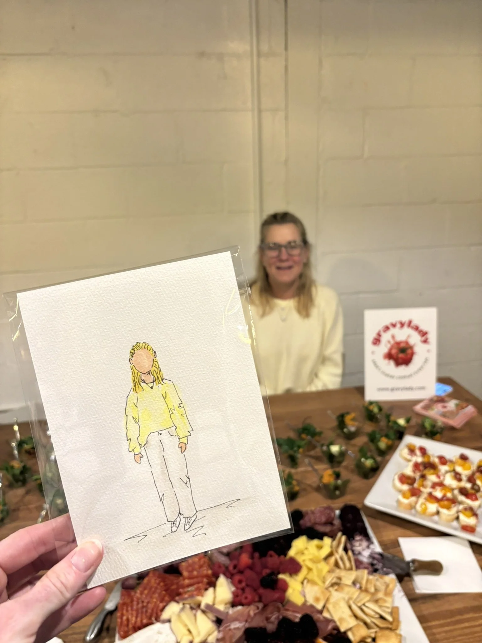 A hand holding a watercolor illustration of a woman in a yellow sweater and light pants, with a blurred woman smiling in the background at a table with various desserts and food items.