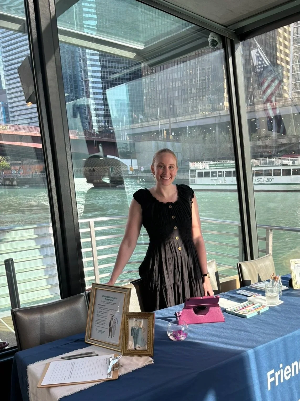 A woman in a black dress smiling, standing behind a table set up with frames, a pink item, and a glass bowl with flowers, inside a glass boat with a view of city buildings and water.