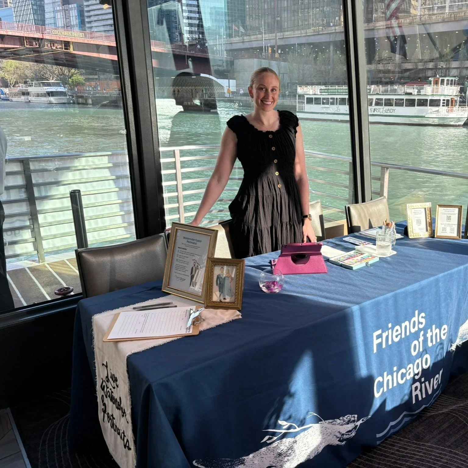 A woman standing behind a table set up for an event, with a city river and boats visible through large windows in the background. The table has various framed pictures, informational materials, and a pink tablet holder.