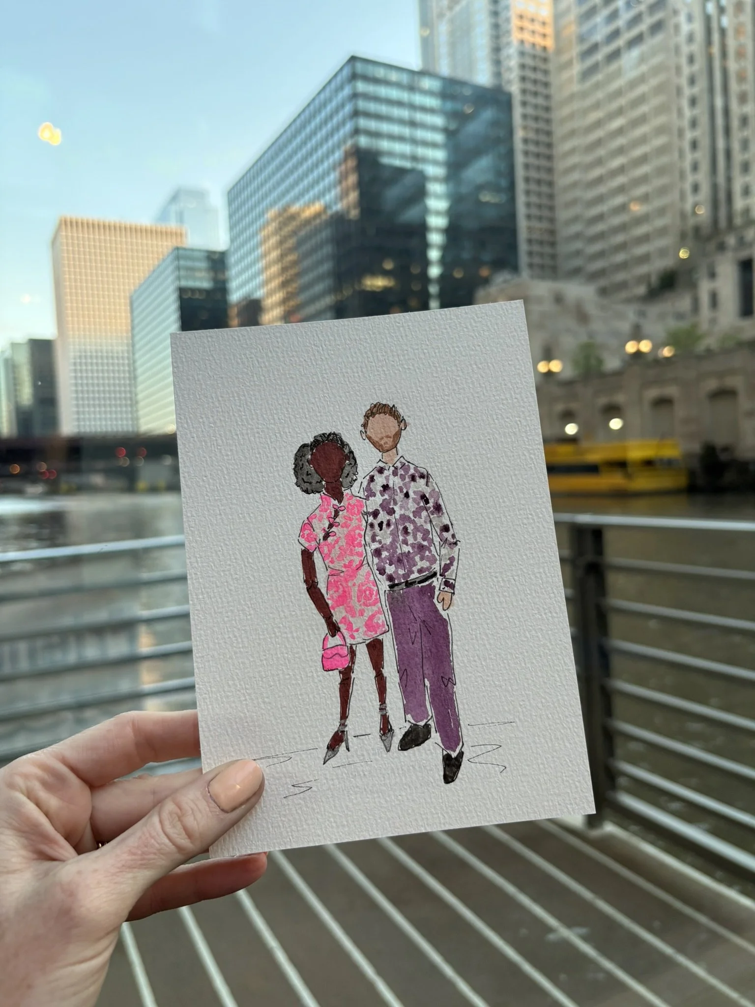 A hand holding a watercolor illustration of two people walking together against a cityscape background with tall glass buildings and a yellow bus.