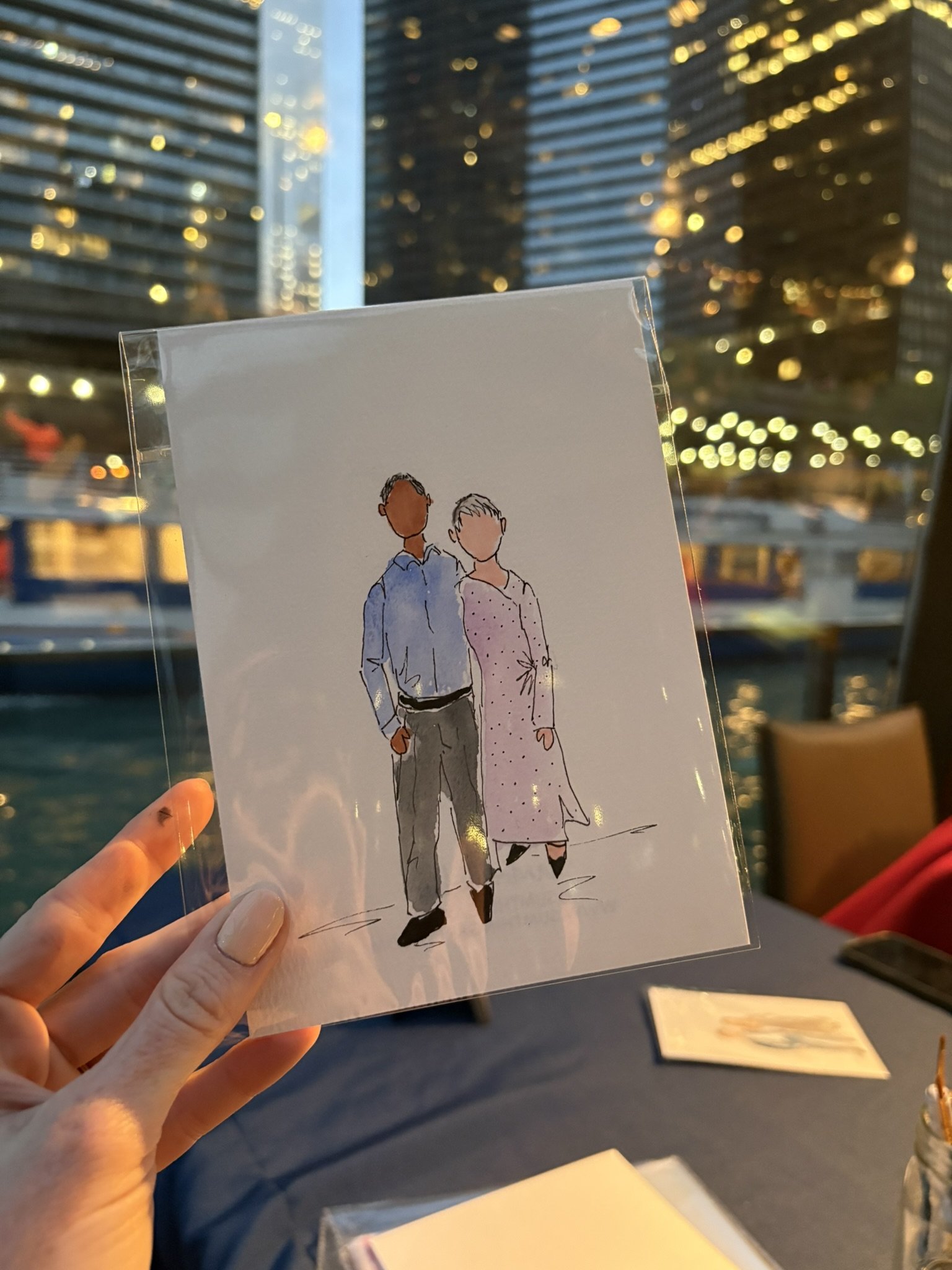 Hand holding a plastic sleeve with a hand-drawn illustration of a man and woman standing side by side, with tall modern buildings and city lights in the background during evening or night.