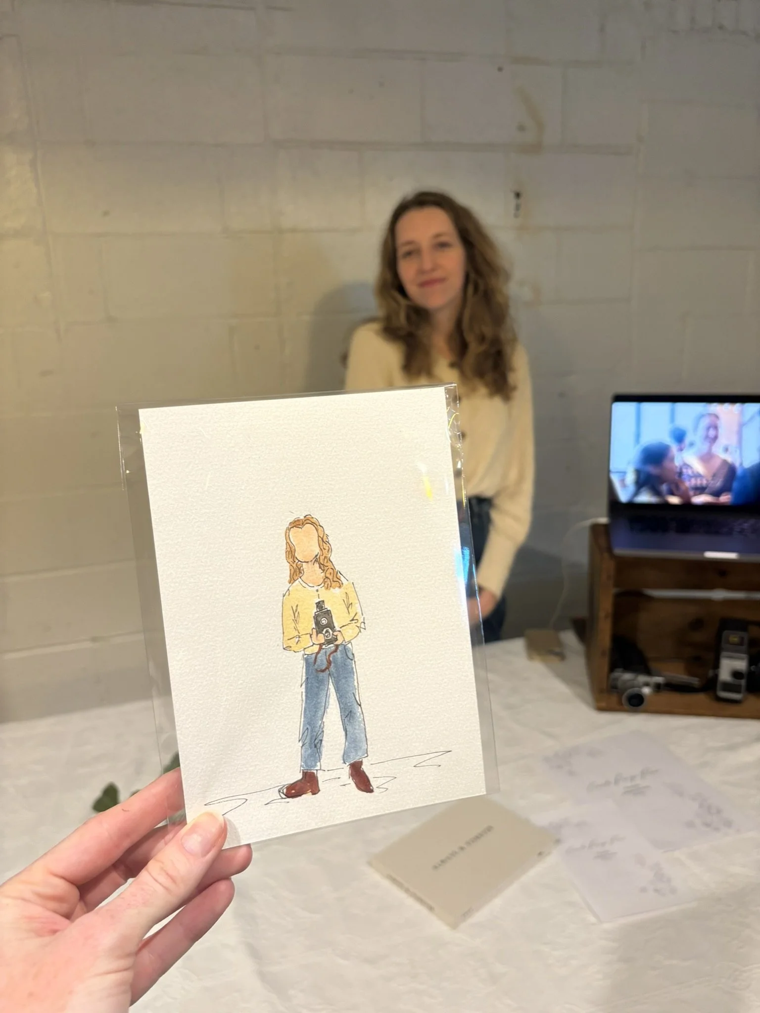 A person holding a small piece of art featuring a woman with curly red hair wearing a beige sweater and blue jeans, holding a camera.