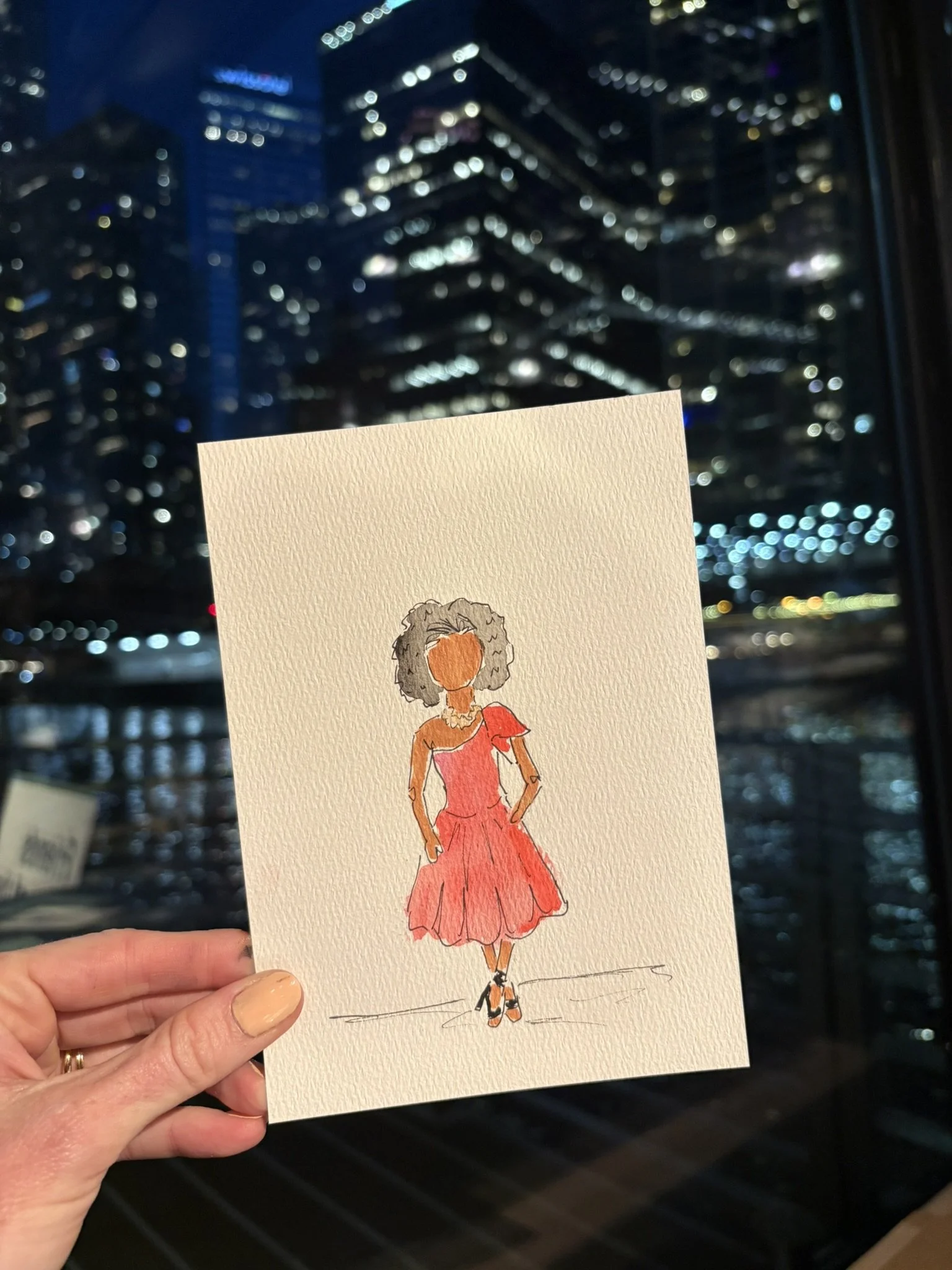 Watercolor fashion illustration of a woman with curly hair wearing an orange dress and matching shoes, held up against a backdrop of city buildings at night.