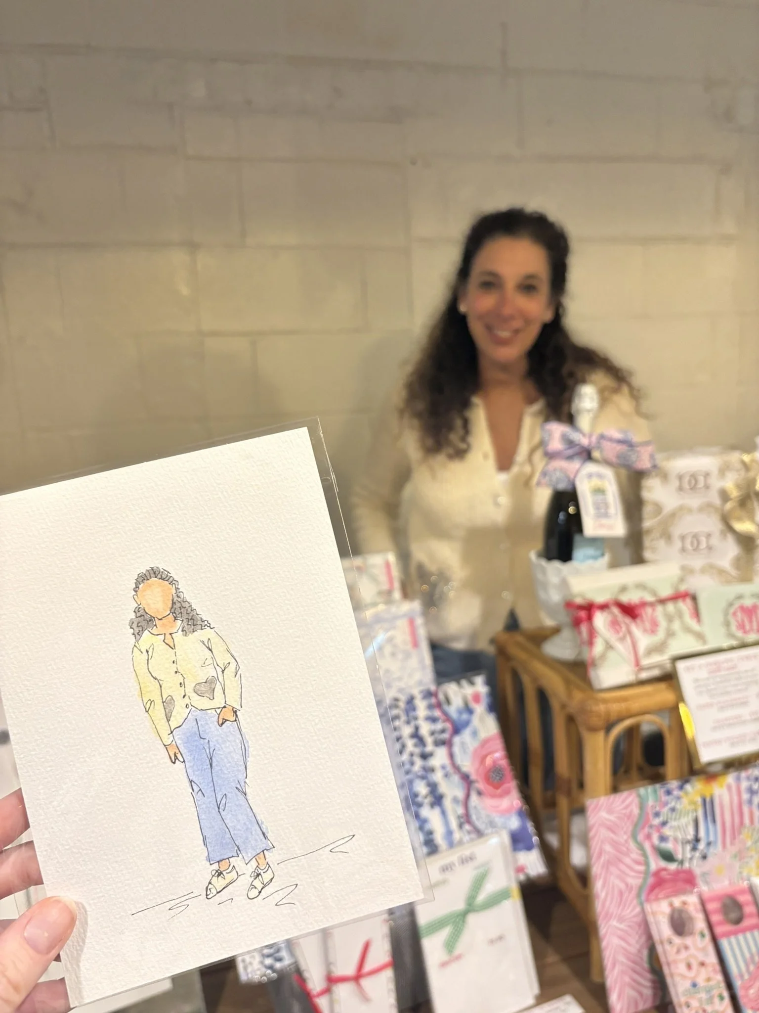 A woman with dark curly hair smiling at a market stall with colorful gift bags and a framed illustration of a woman in a yellow jacket and jeans.