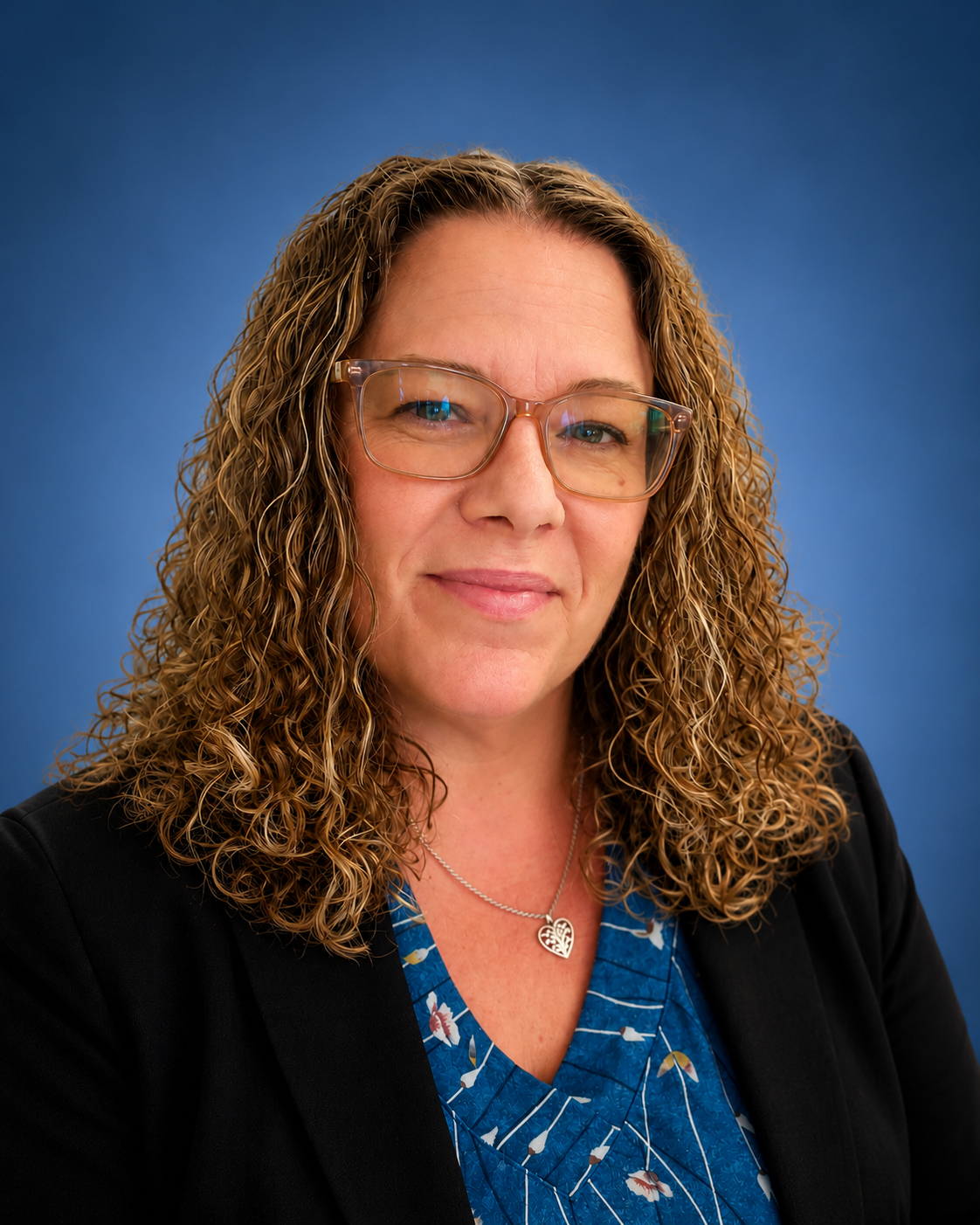 A woman with curly blonde hair wearing glasses, a black blazer, a blue patterned blouse, and a silver heart necklace, posing against a blue backgroundCare Management Green Valley AZ