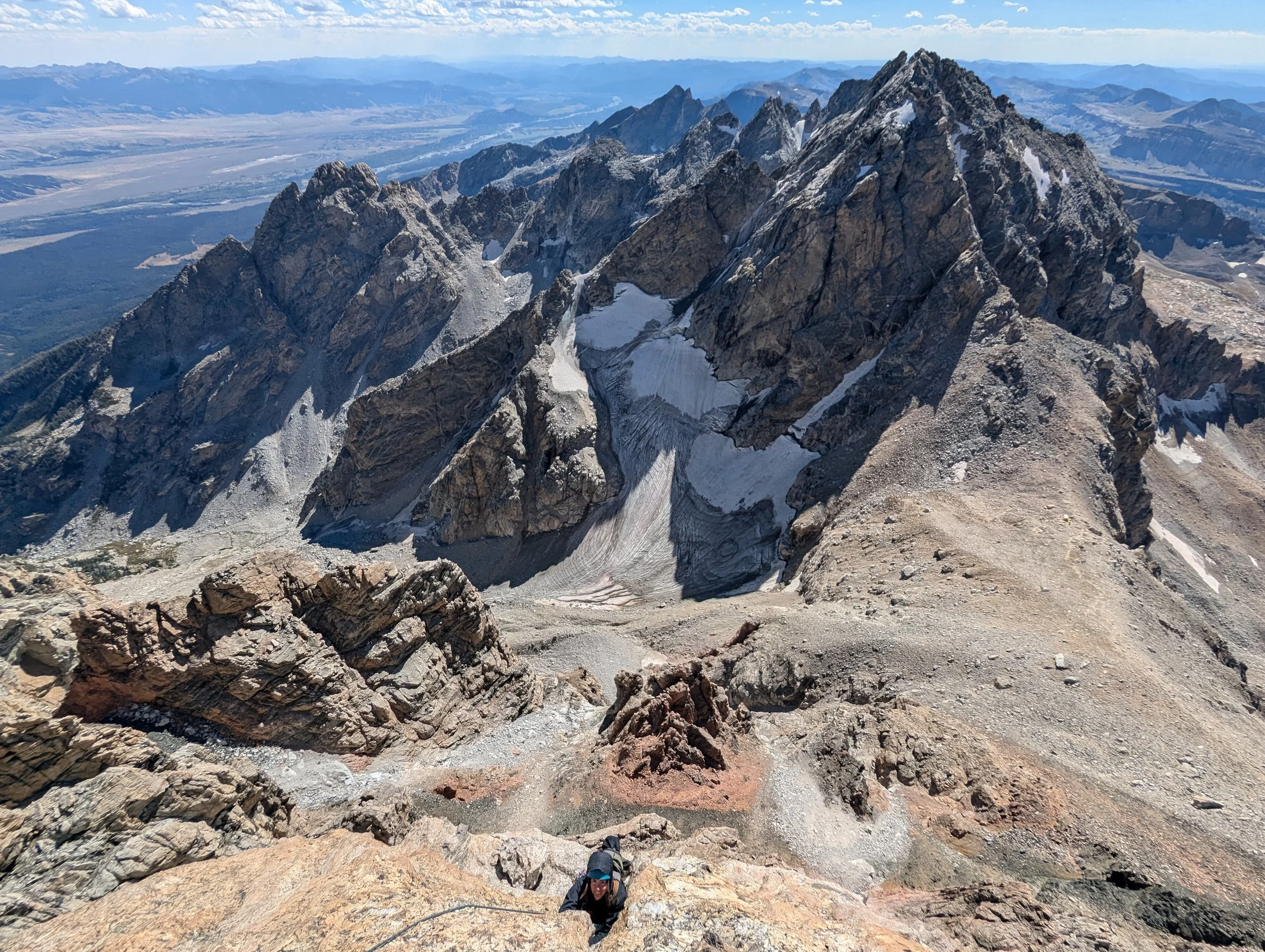 Climber ascending a rugged mountain slope with a view of snow-capped peaks and a valley in the distance.