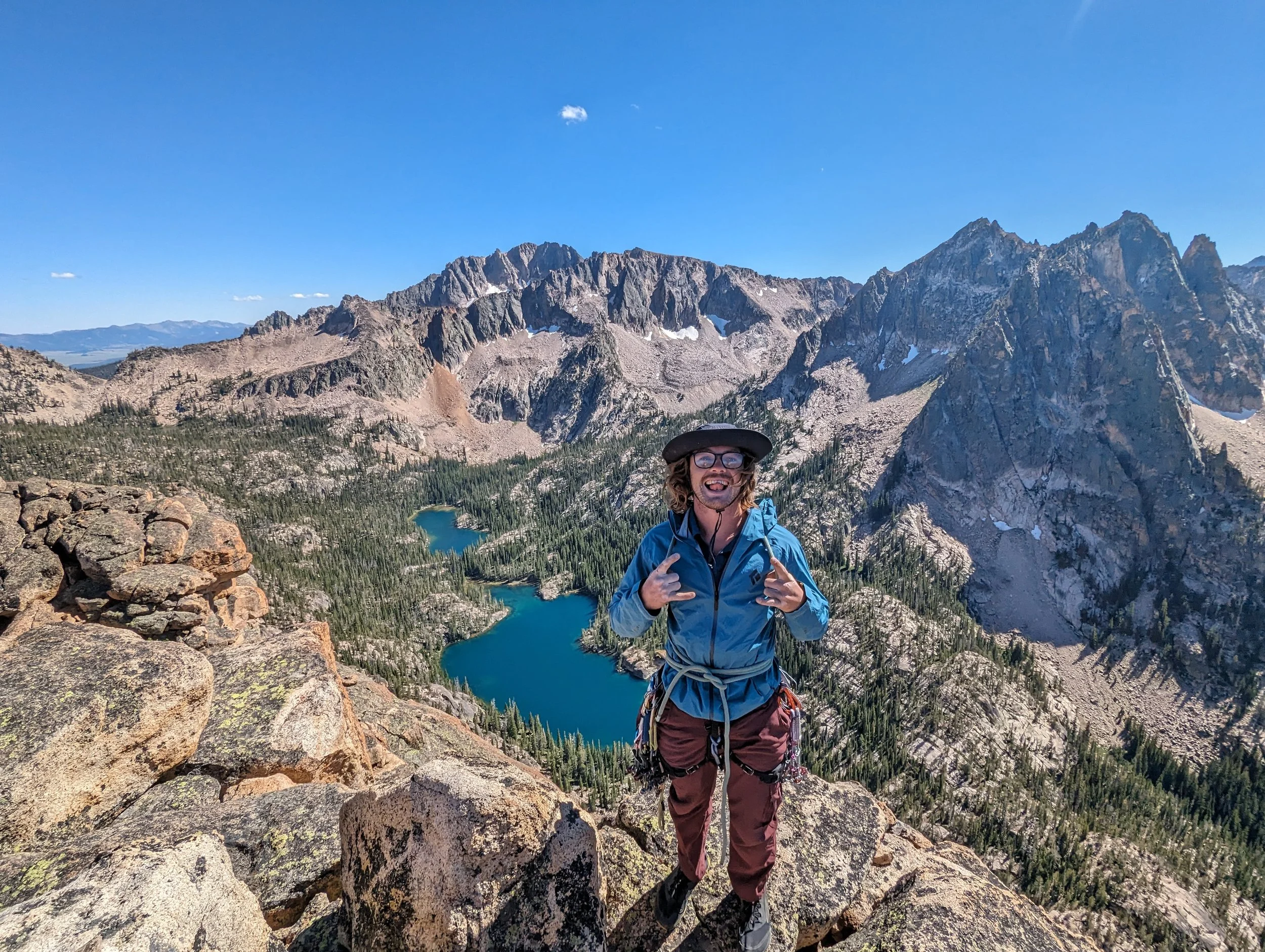 A person standing on rocks in a mountainous landscape with a lake and evergreen trees, under a clear blue sky.