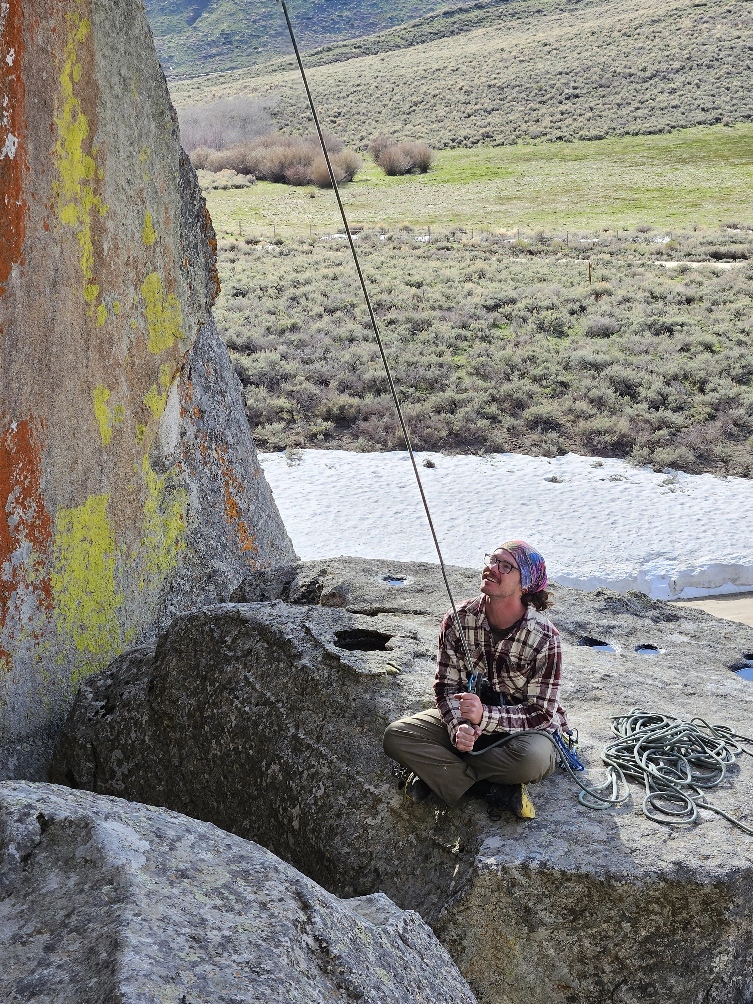Person rock climbing with a sling and rope on a large boulder outdoors in a mountainous area, smiling and looking up.