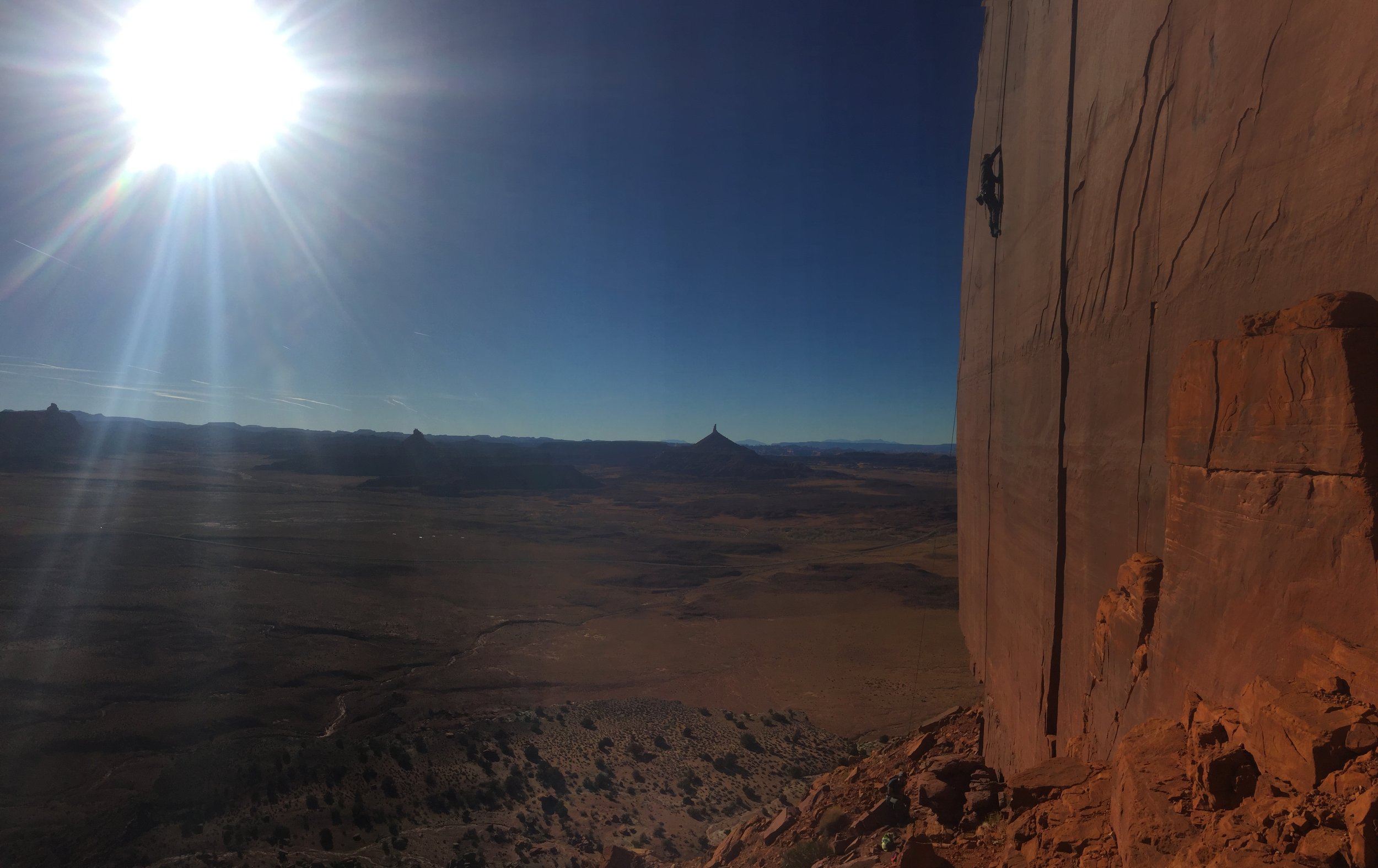A vast desert landscape with a prominent flat-topped mesa and a pointed mountain in the distance, viewed from a high vantage point. The sky is clear and bright with the sun shining intensely.