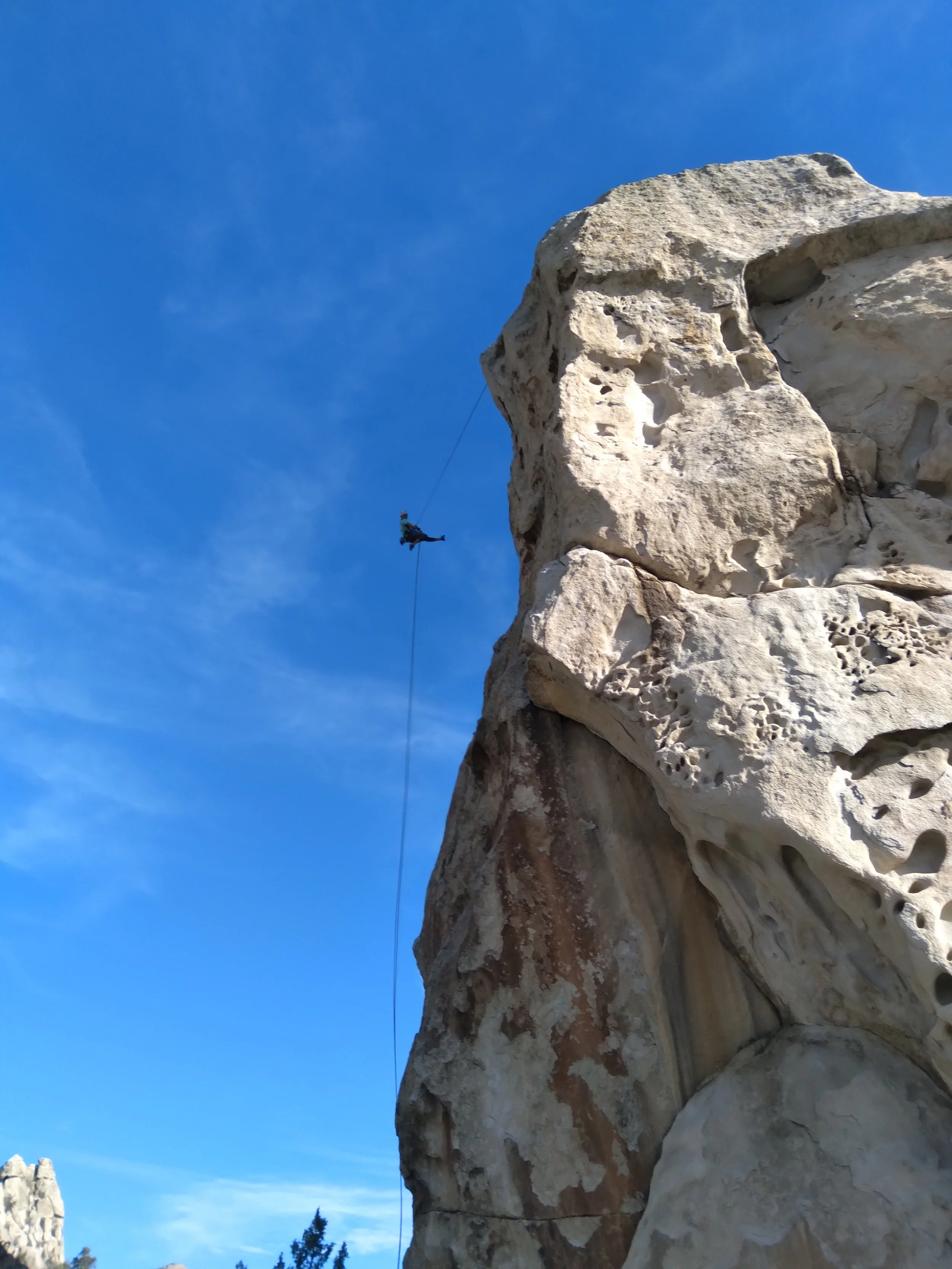 A person climbing a rock formation with a harness, hanging from a rope against a clear blue sky.
