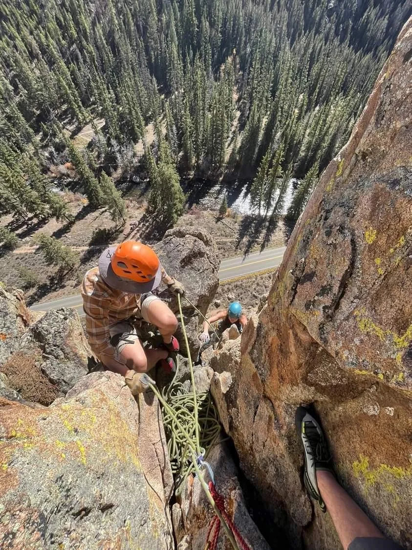 Climbers ascending a steep rock face or cliff with ropes, views of a forested landscape below.