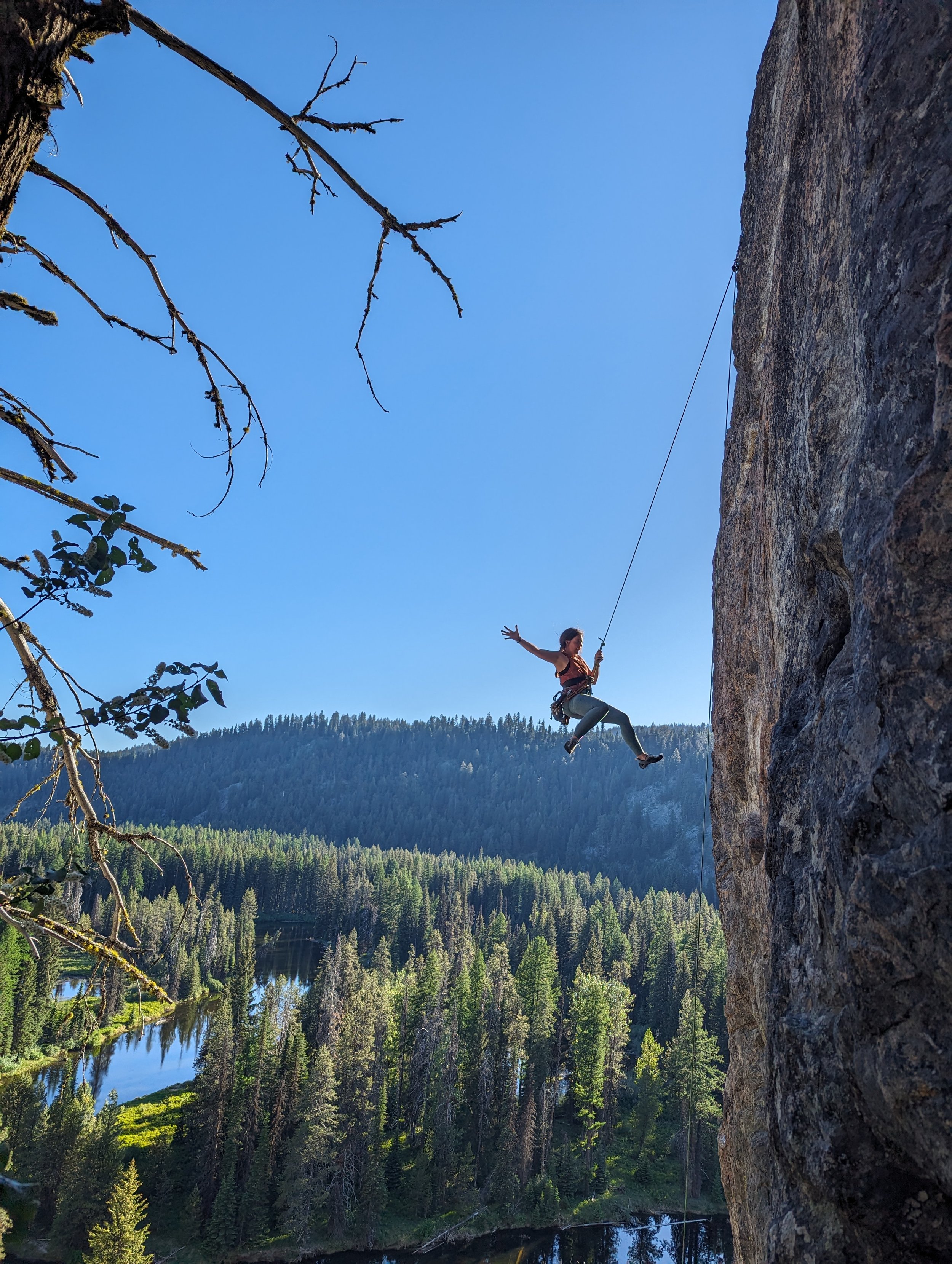 A woman is rock climbing outdoors with a harness, while holding a rope, with a forest and lake below her and a clear blue sky above.