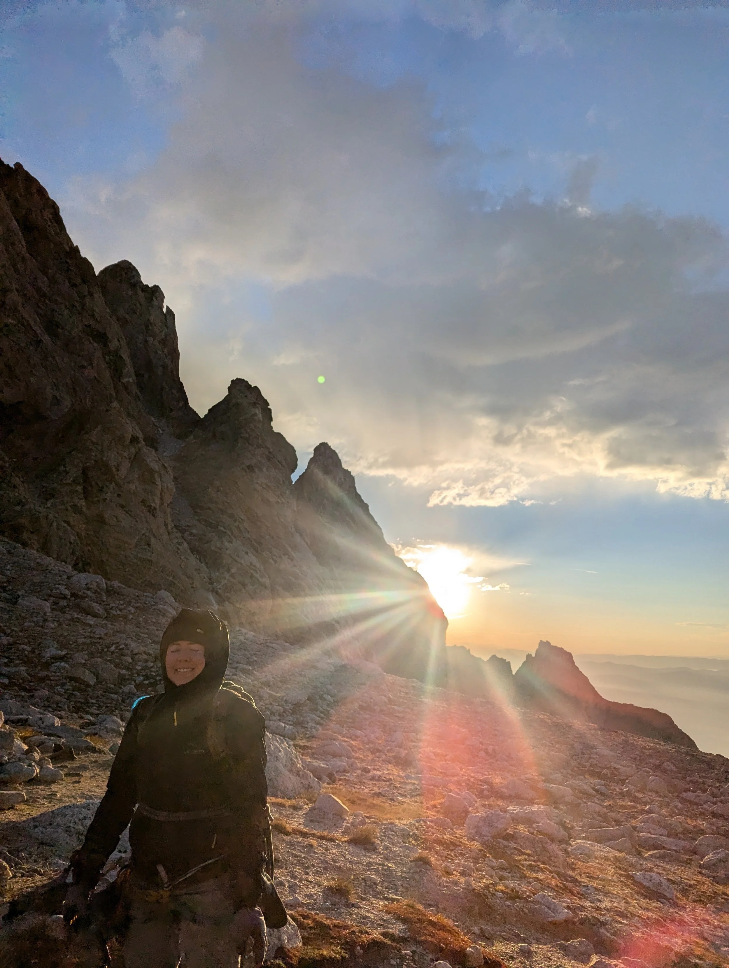 A smiling person with a backpack standing on rocky terrain with mountains, clouds, and the sun setting behind them, creating a sunset glow.