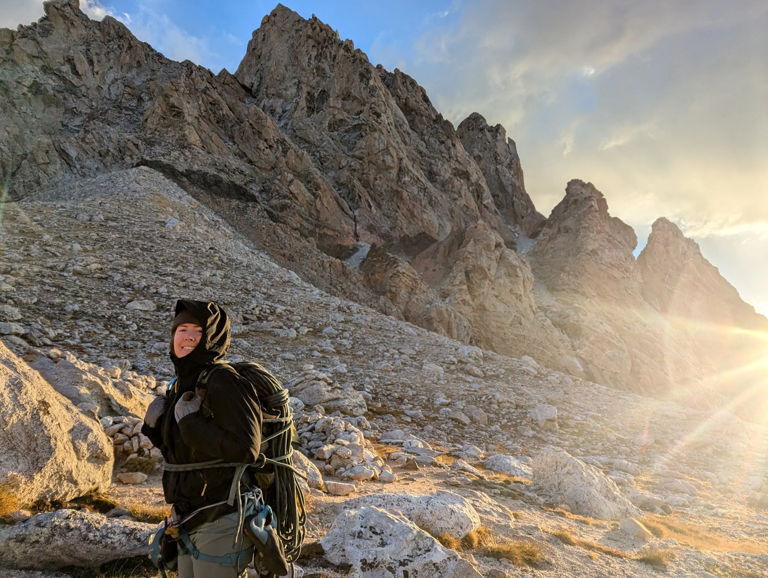 A person dressed in black outdoor clothing with a backpack and climbing gear, standing on rocky terrain near a mountain with steep rocky cliffs, during sunrise or sunset.