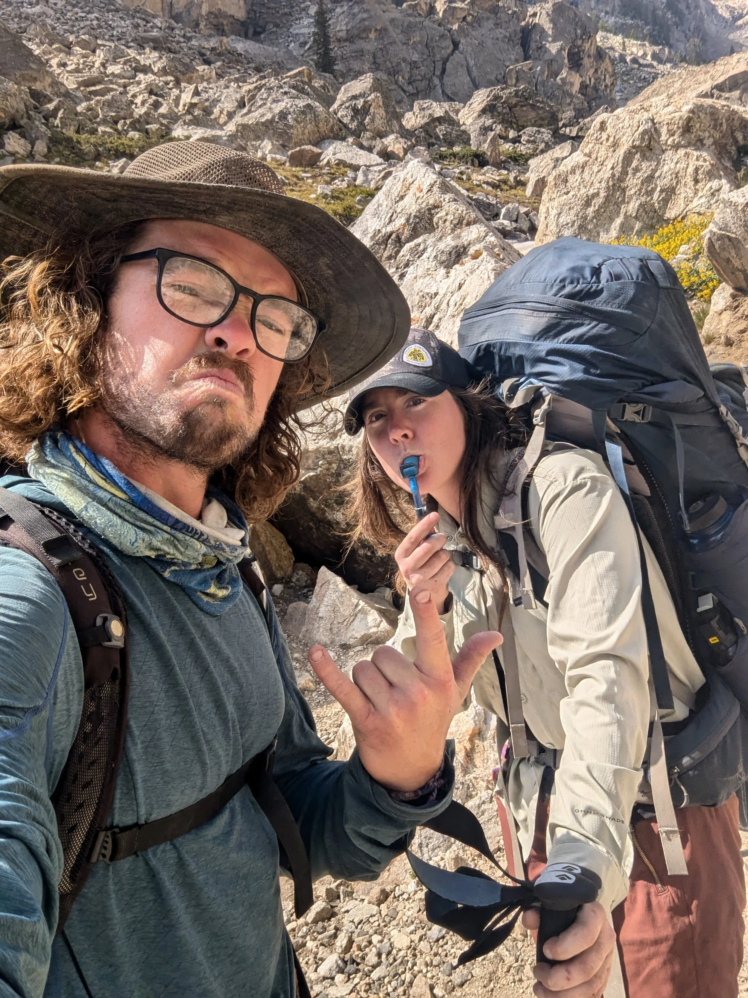 Two hikers in rocky mountain terrain, one man with a large hat and glasses taking a selfie, and a woman with a backpack and a whistle making a hand sign.