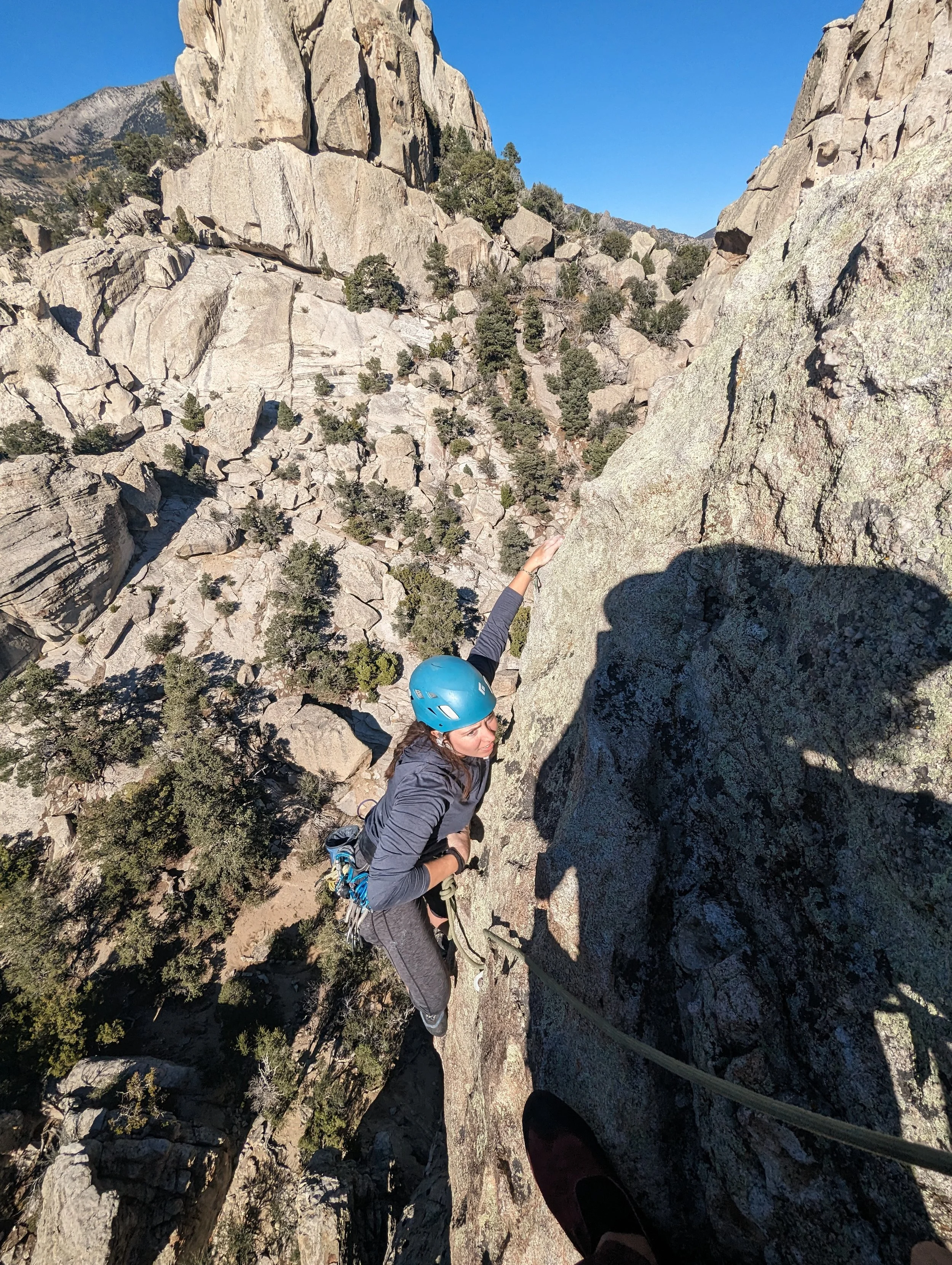 A person in a blue helmet rock climbing on a rugged, rocky mountain face with trees and distant mountains in the background.