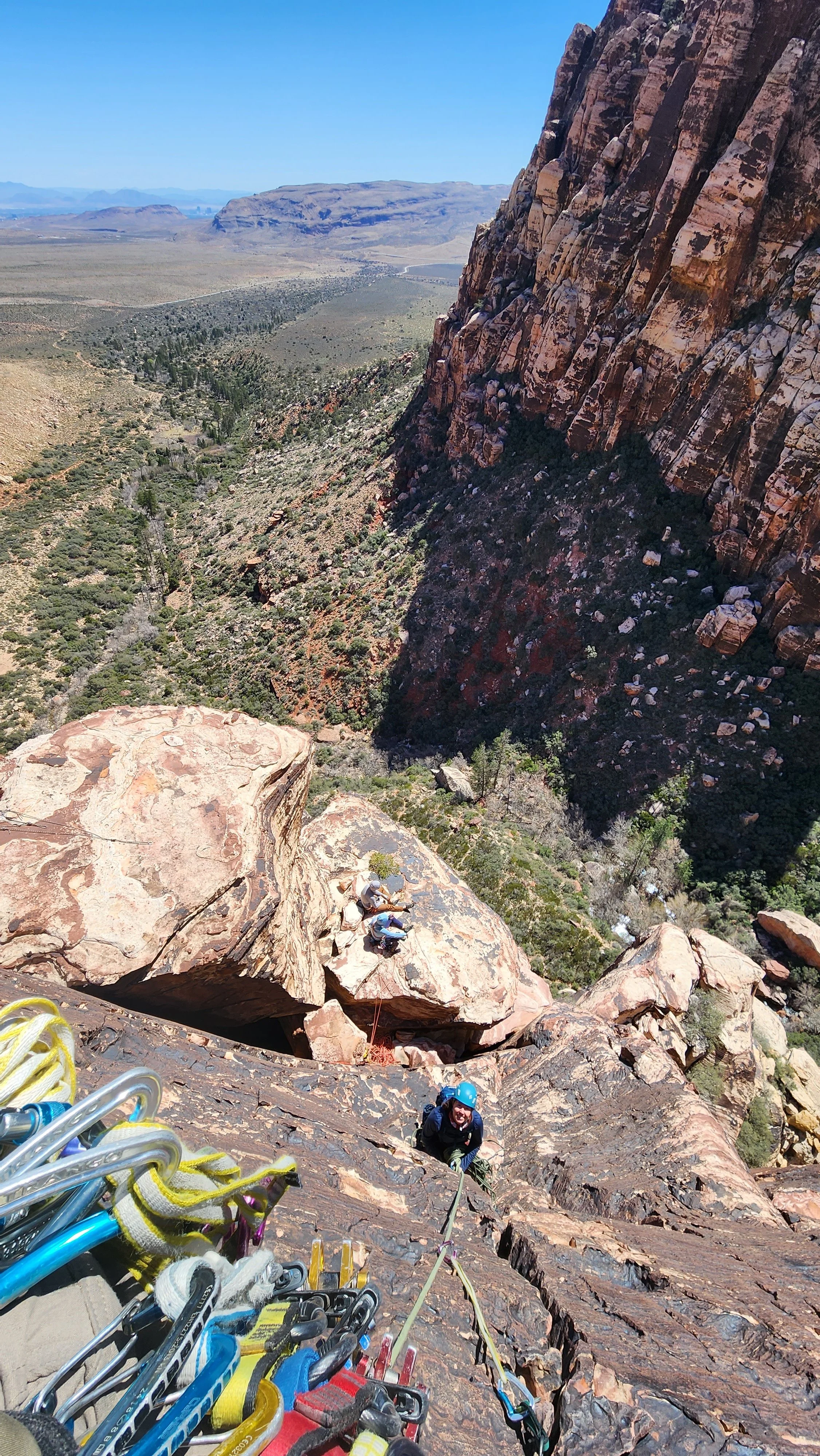 View from a rock climbing route showing climbers on red rock formation, with vast desert landscape and mountains in the background.