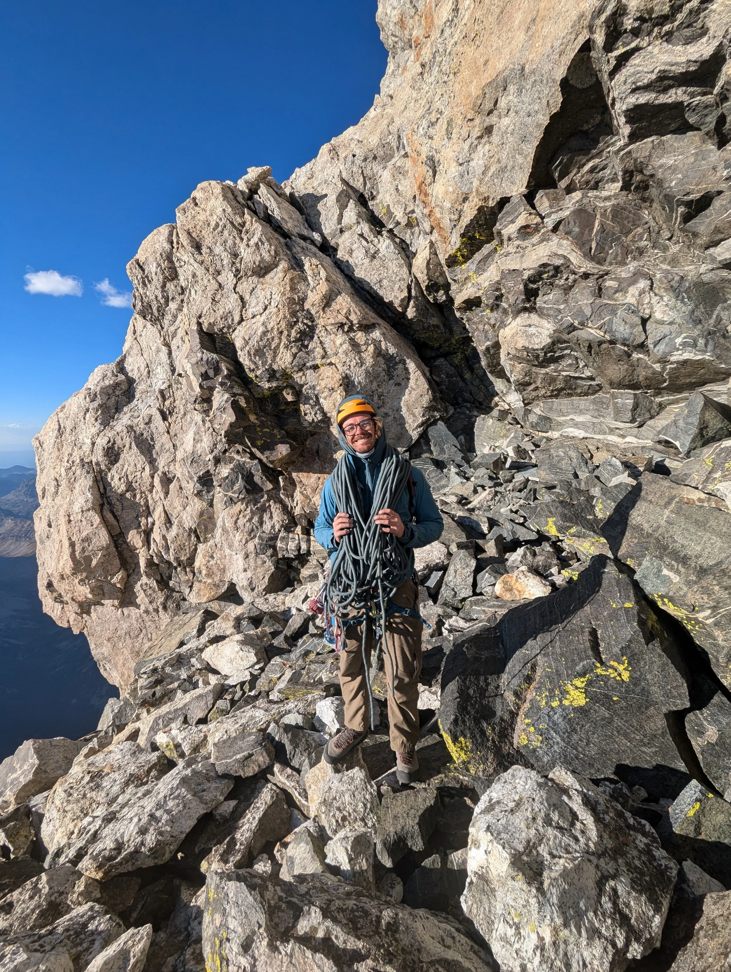 A smiling male climber standing on a rocky mountain ledge, holding climbing gear and wearing a helmet, with steep rocky cliffs behind him and a clear blue sky overhead.