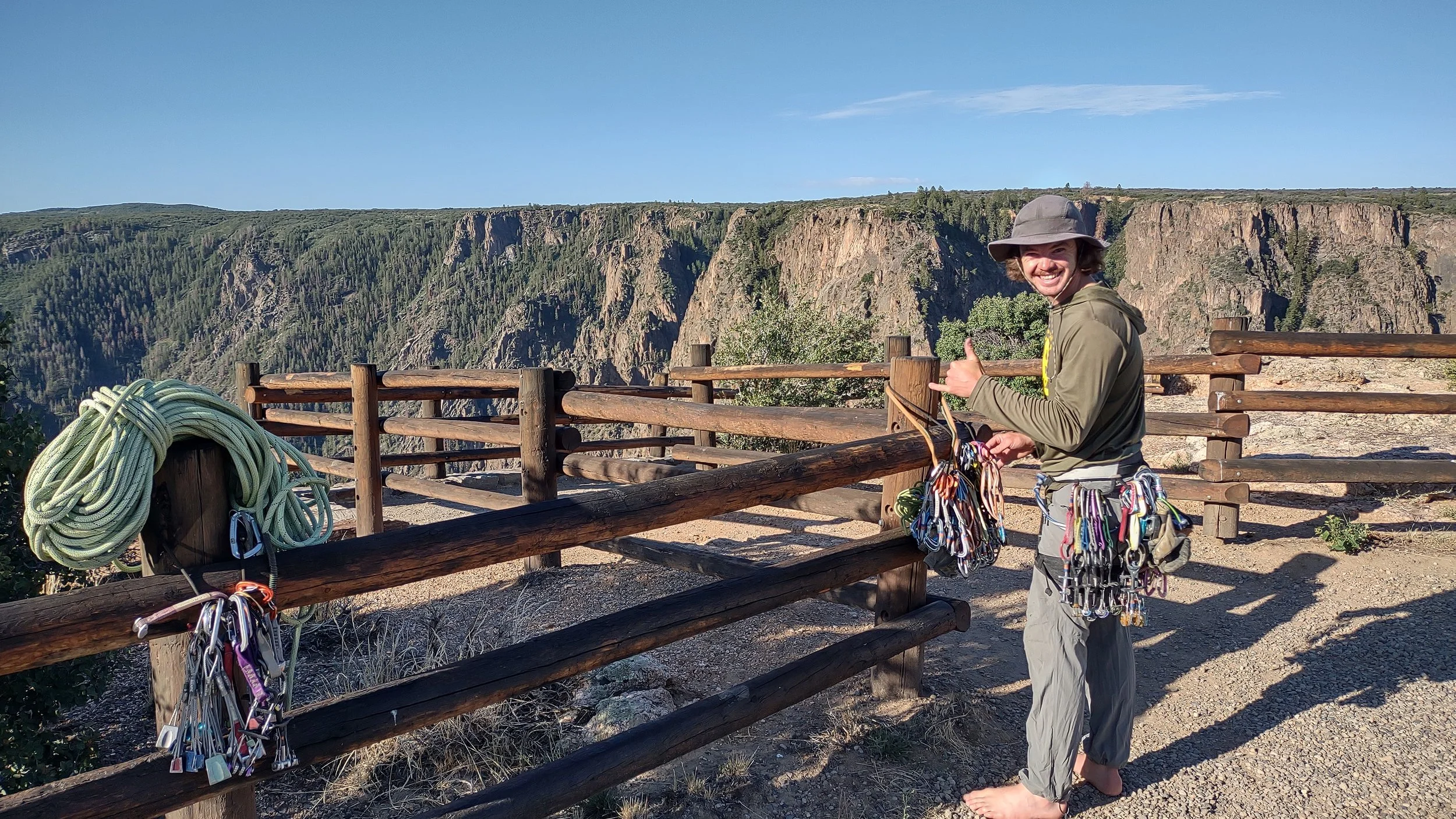 A smiling climber giving a thumbs up standing behind a wooden fence on a rocky overlook with a canyon and forested hills in the background.
