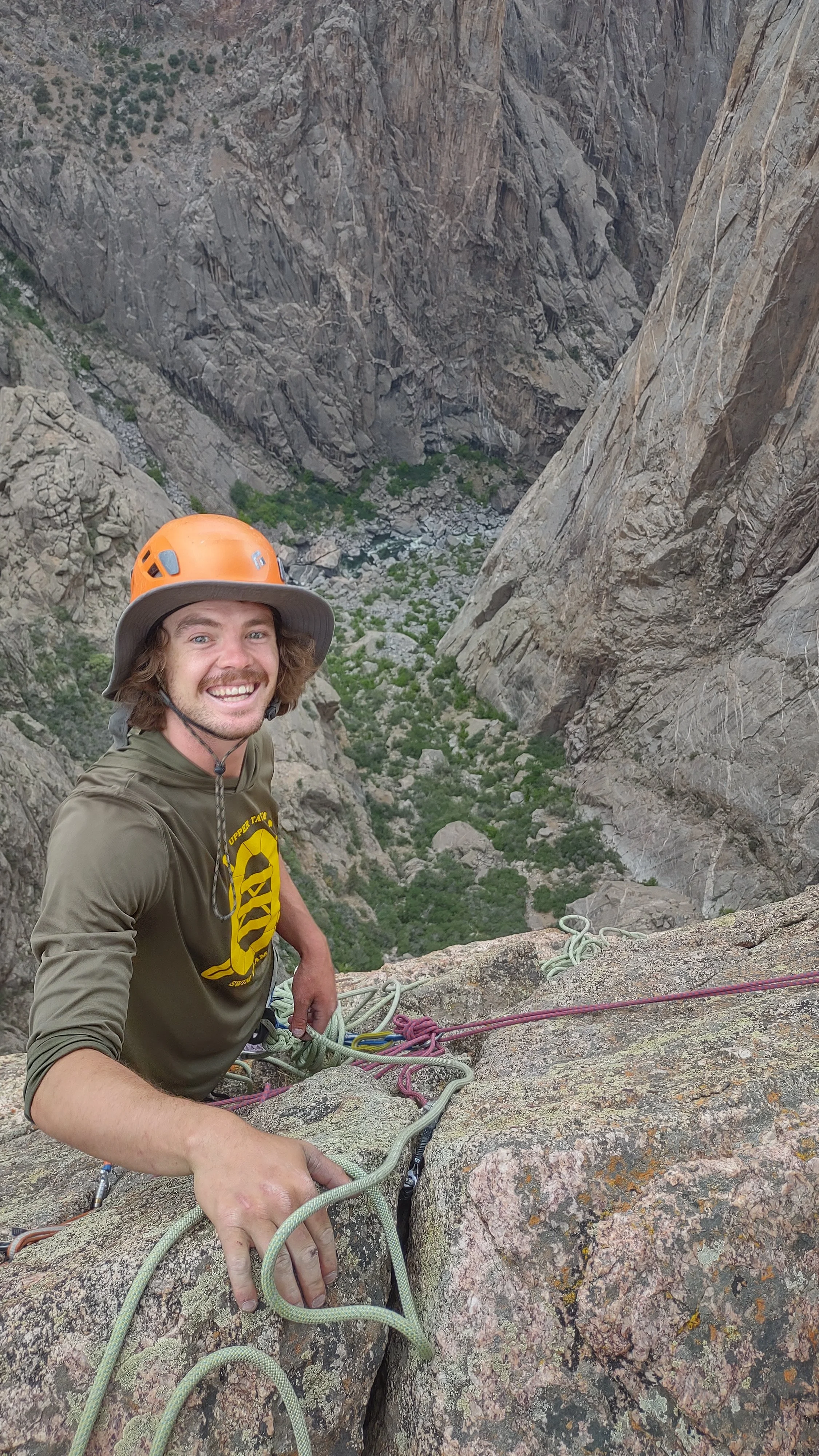 A man wearing a safety helmet and climbing gear, smiling while climbing on a rock face in a canyon with steep cliffs and a green valley below.