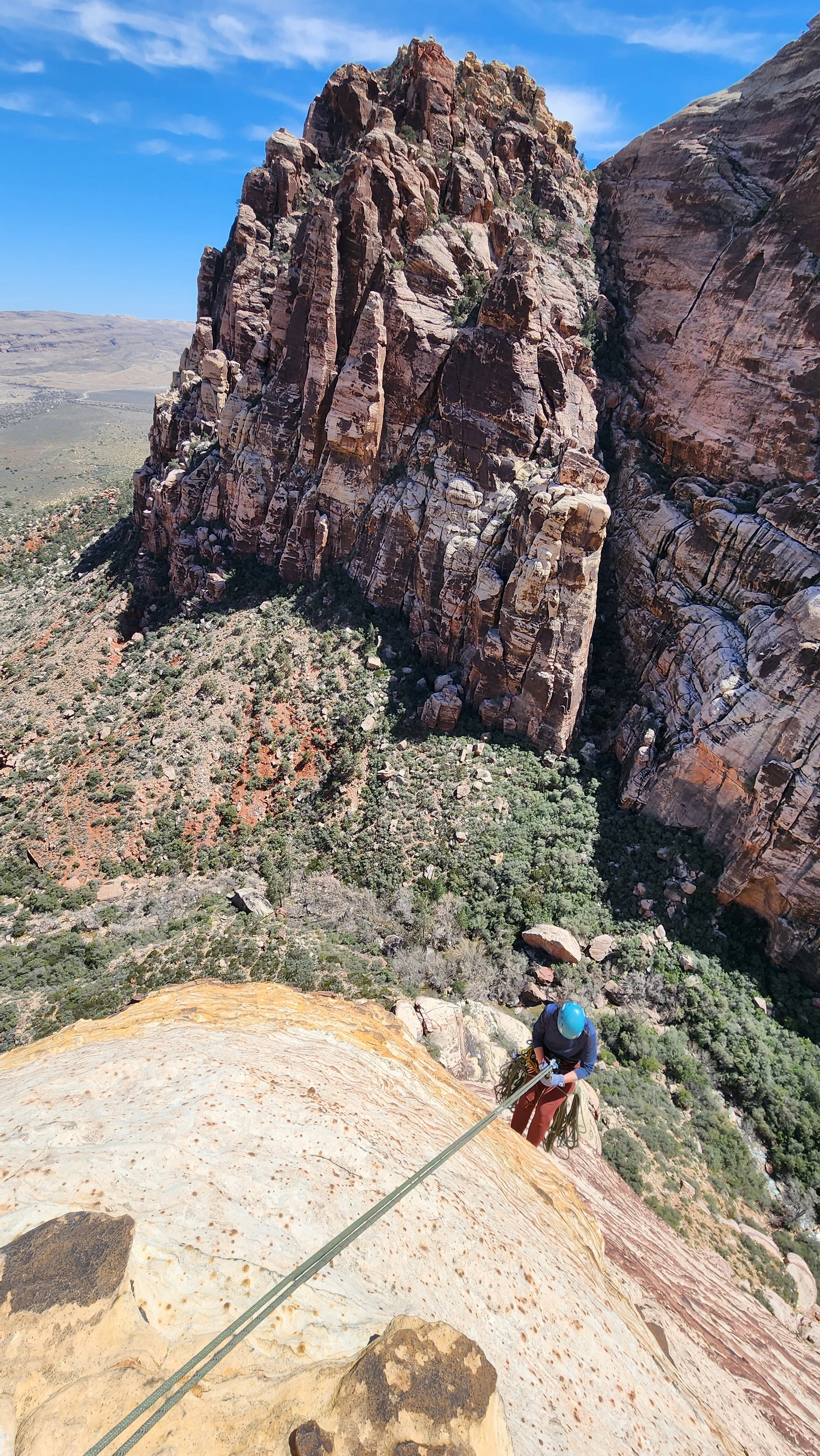 A person wearing a blue helmet and climbing gear rappelling down a rocky cliff face with a large canyon and rugged terrain in the background.