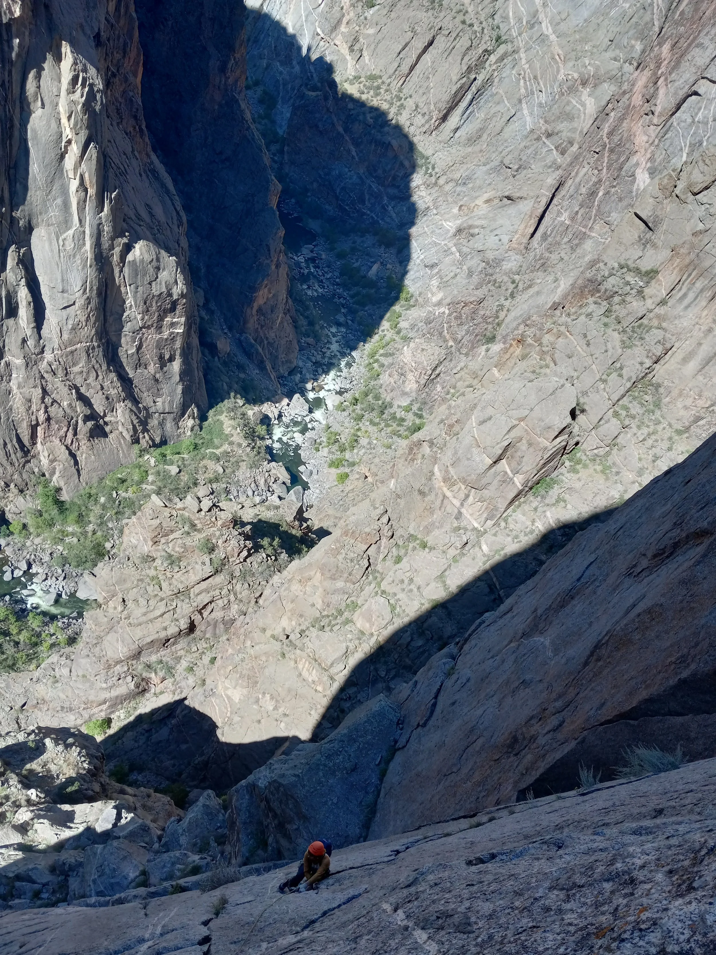 A person wearing a helmet and climbing gear is ascending a steep rock face in a deep canyon with towering vertical rock walls and a river at the bottom.