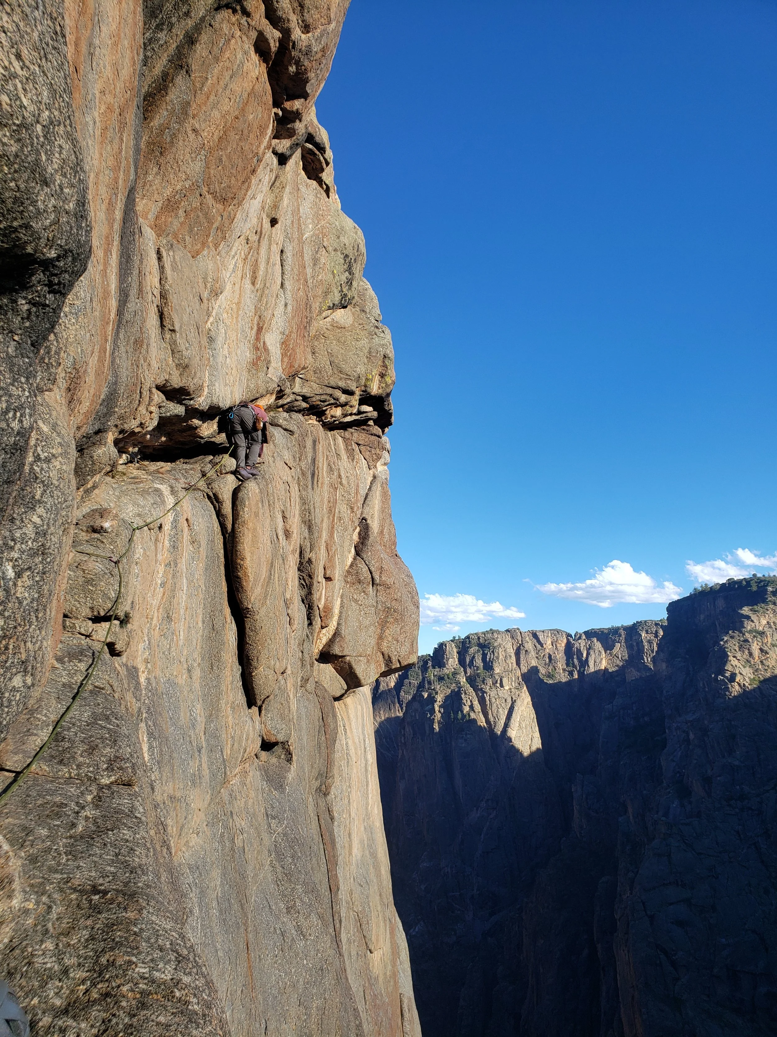 A person rock climbing on a steep cliff face with a safety rope, overlooking a canyon landscape with mountains and a clear blue sky.