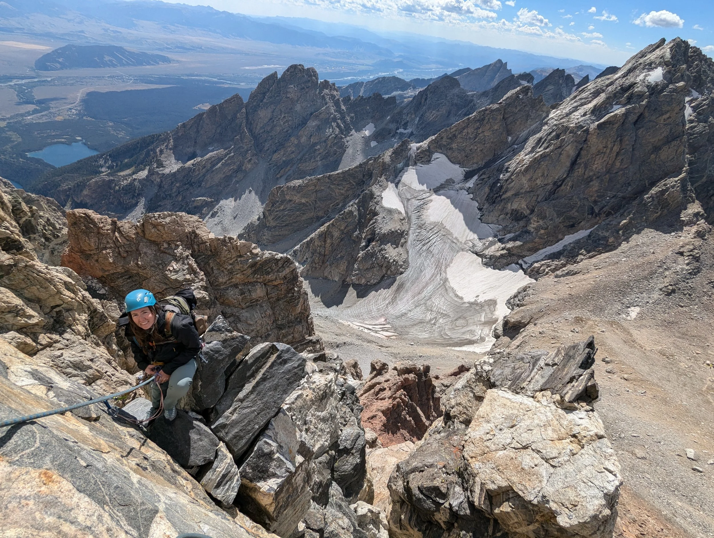 A woman wearing a helmet and climbing gear ascends on a rocky mountain trail with a scenic mountain landscape and glaciers in the background.