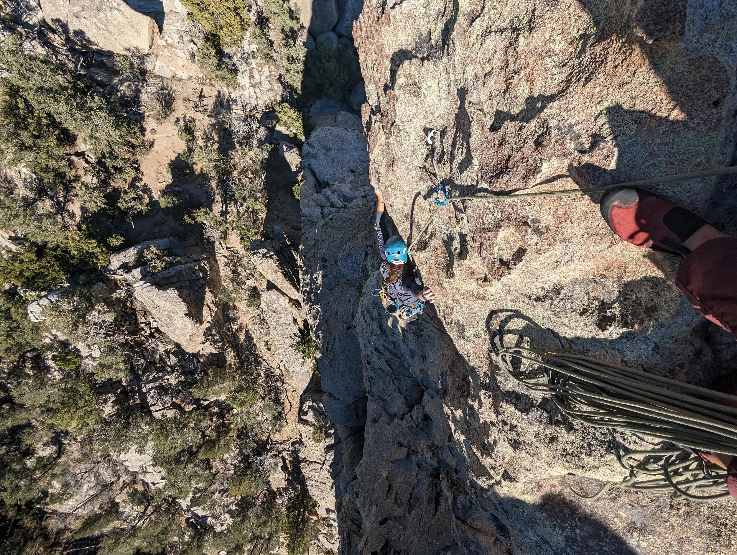 A person rock climbing on a steep cliff face, wearing a blue helmet and black climbing gear, with the perspective from above looking down.