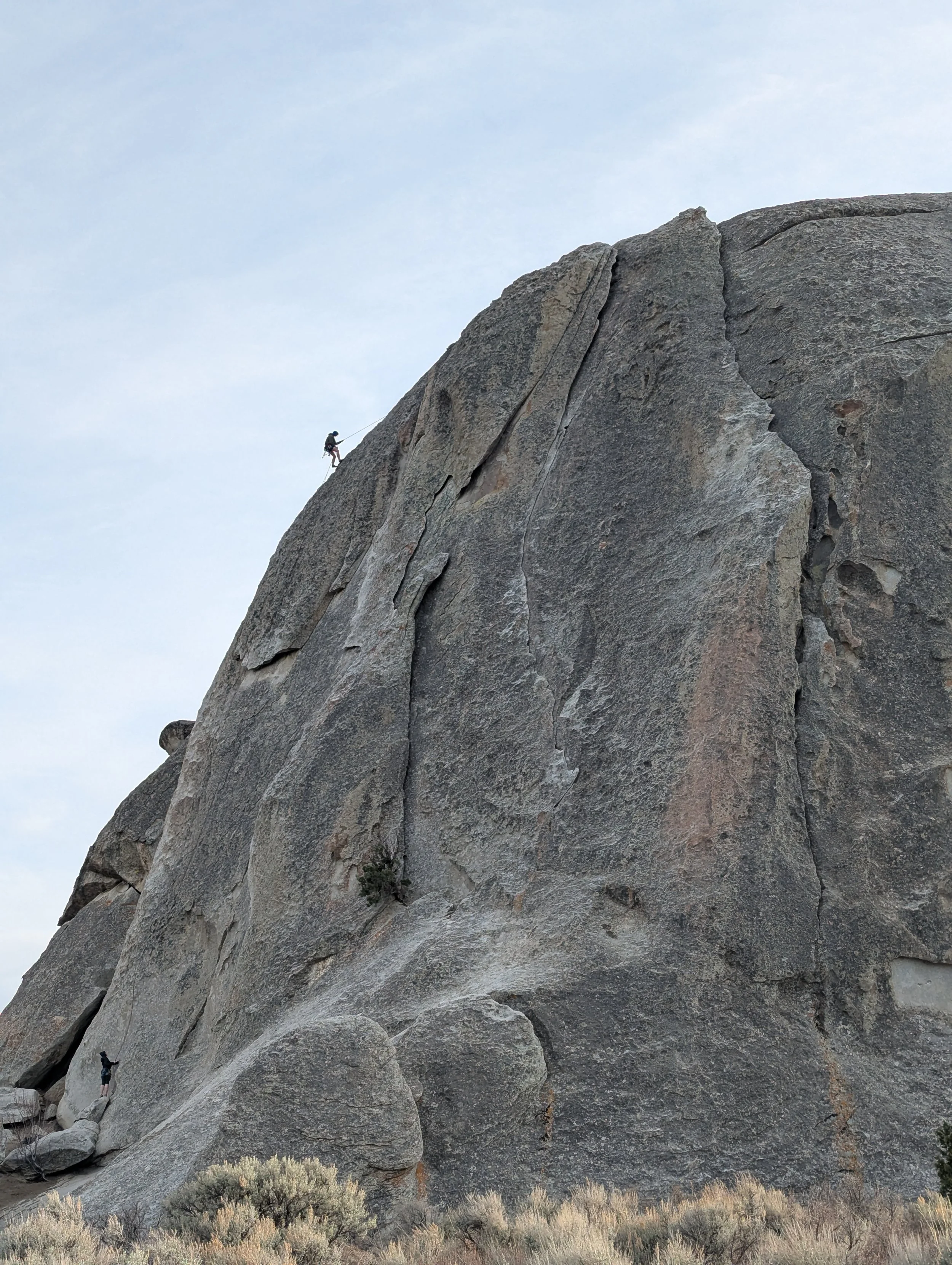 A person rock climbing on a large granite mountain, with another climber at the bottom, surrounded by desert vegetation under a cloudy sky.