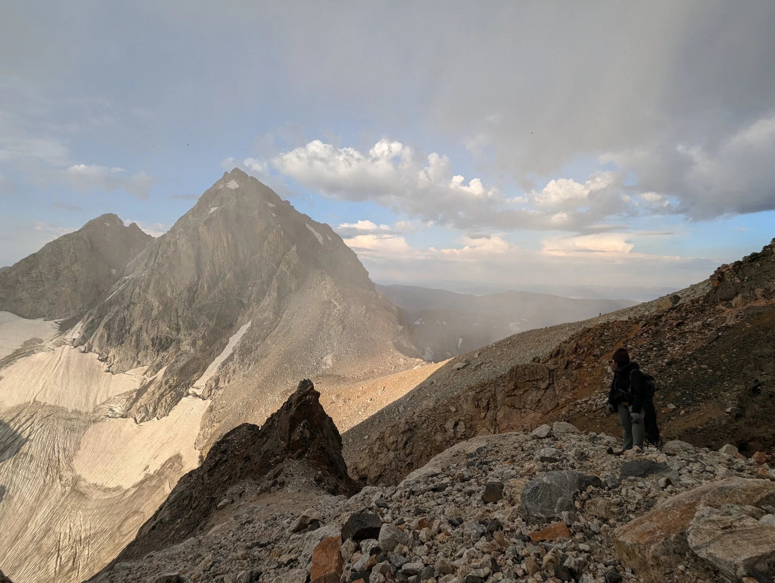 A hiker standing on rocky terrain on a mountainside, with a large mountain peak and snow patches in the background under a partly cloudy sky.