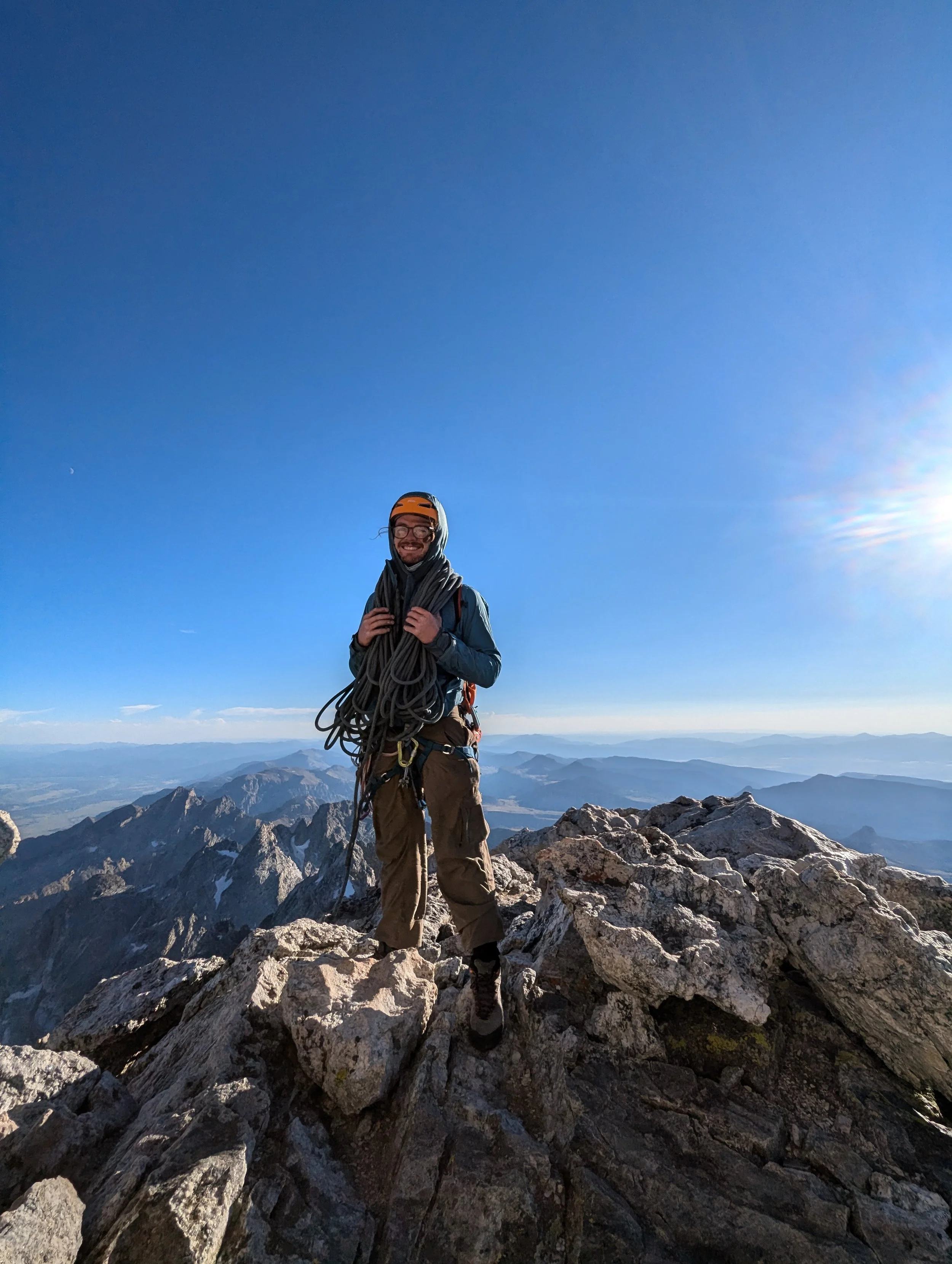 A male climber standing on rocky mountain summit holding climbing rope, with mountain range and clear blue sky in background.