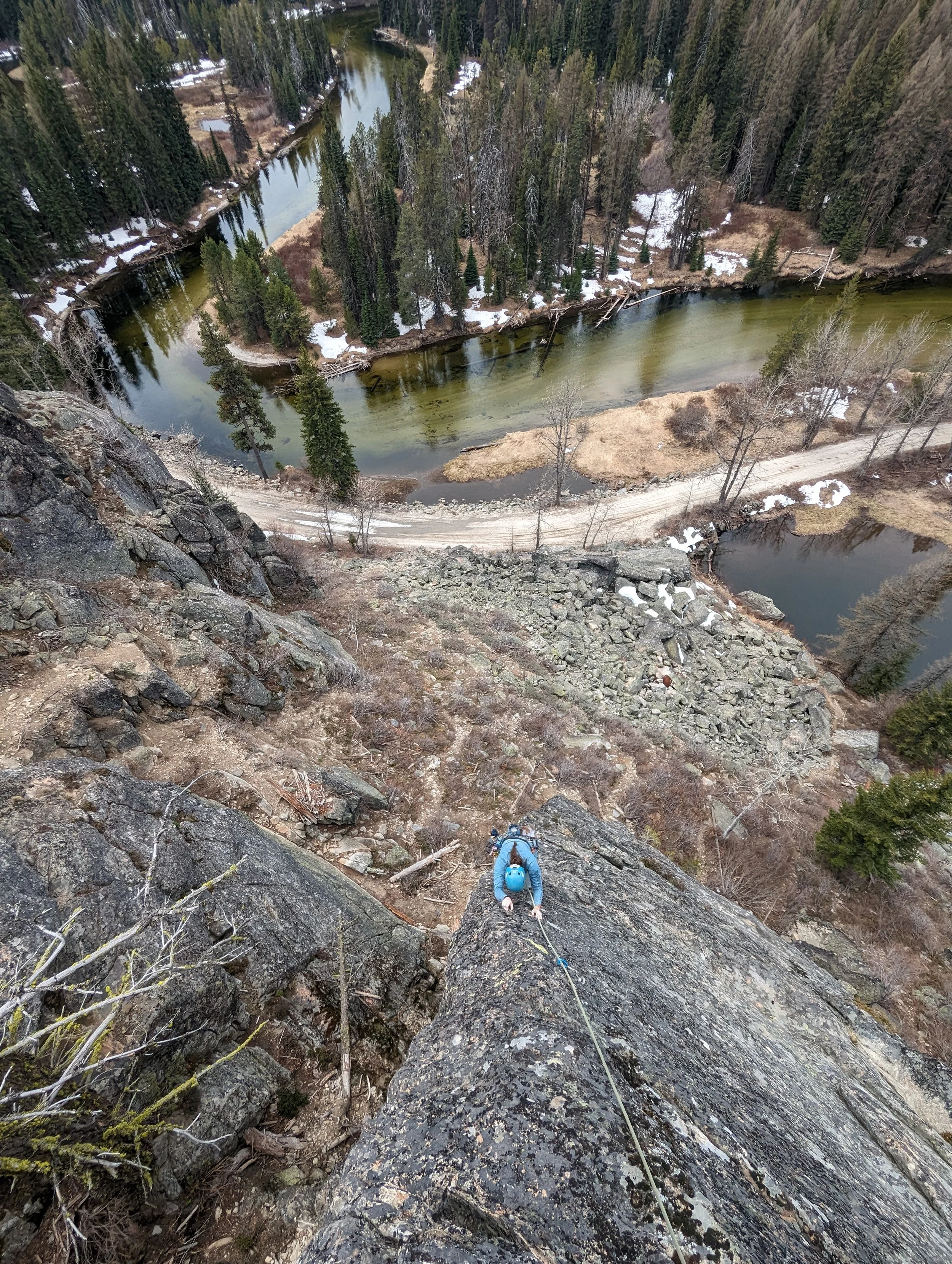 Person climbing a rocky cliff with a rope in a forested area near a river, with trees and patches of snow visible.