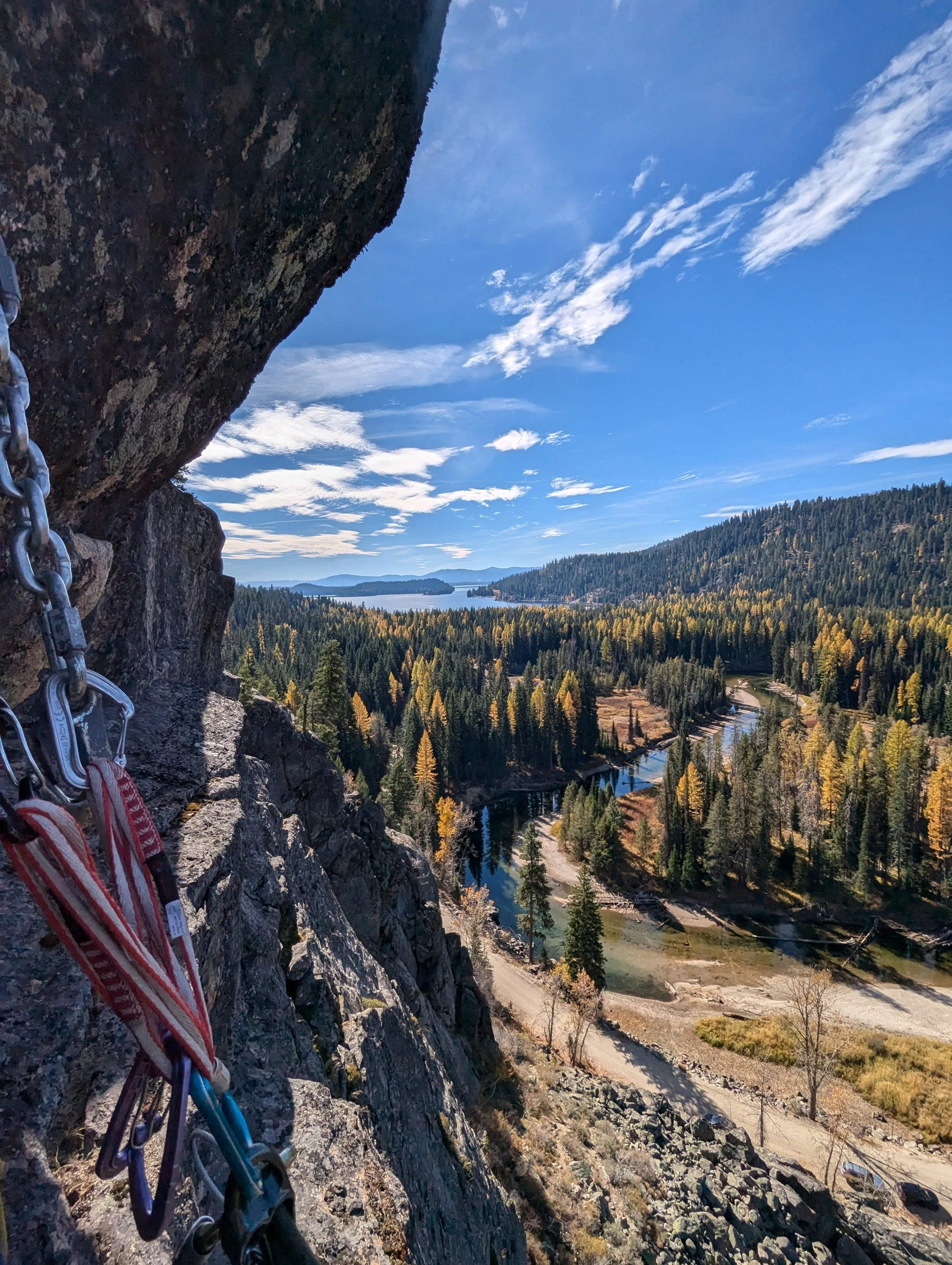 View from a mountain cliff overlooking a forested valley, river, and distant mountains with a blue sky and clouds.