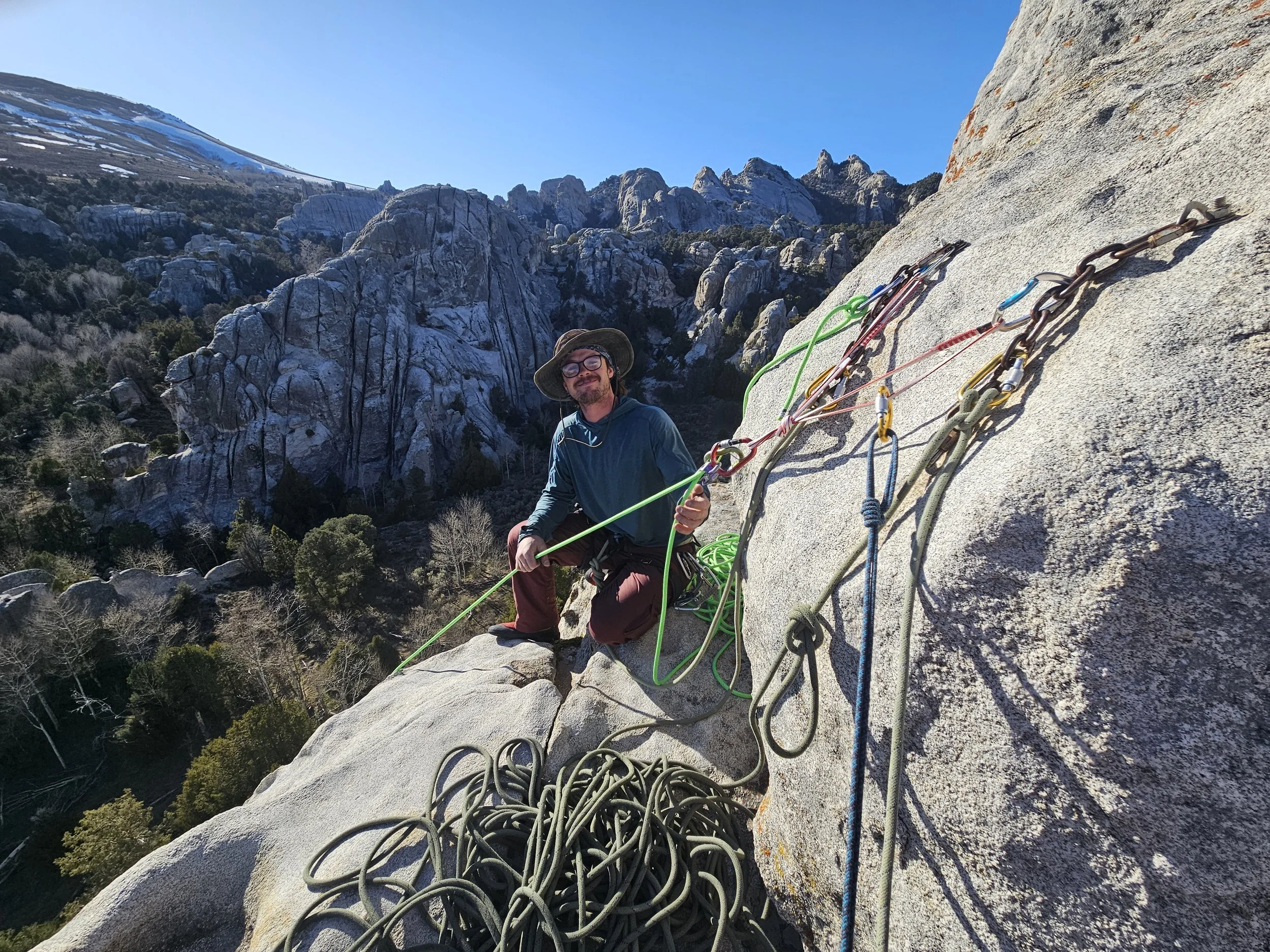 A person in climbing gear is sitting on a rock face overlooking a mountain landscape, with climbing ropes and gear around them on a sunny day.