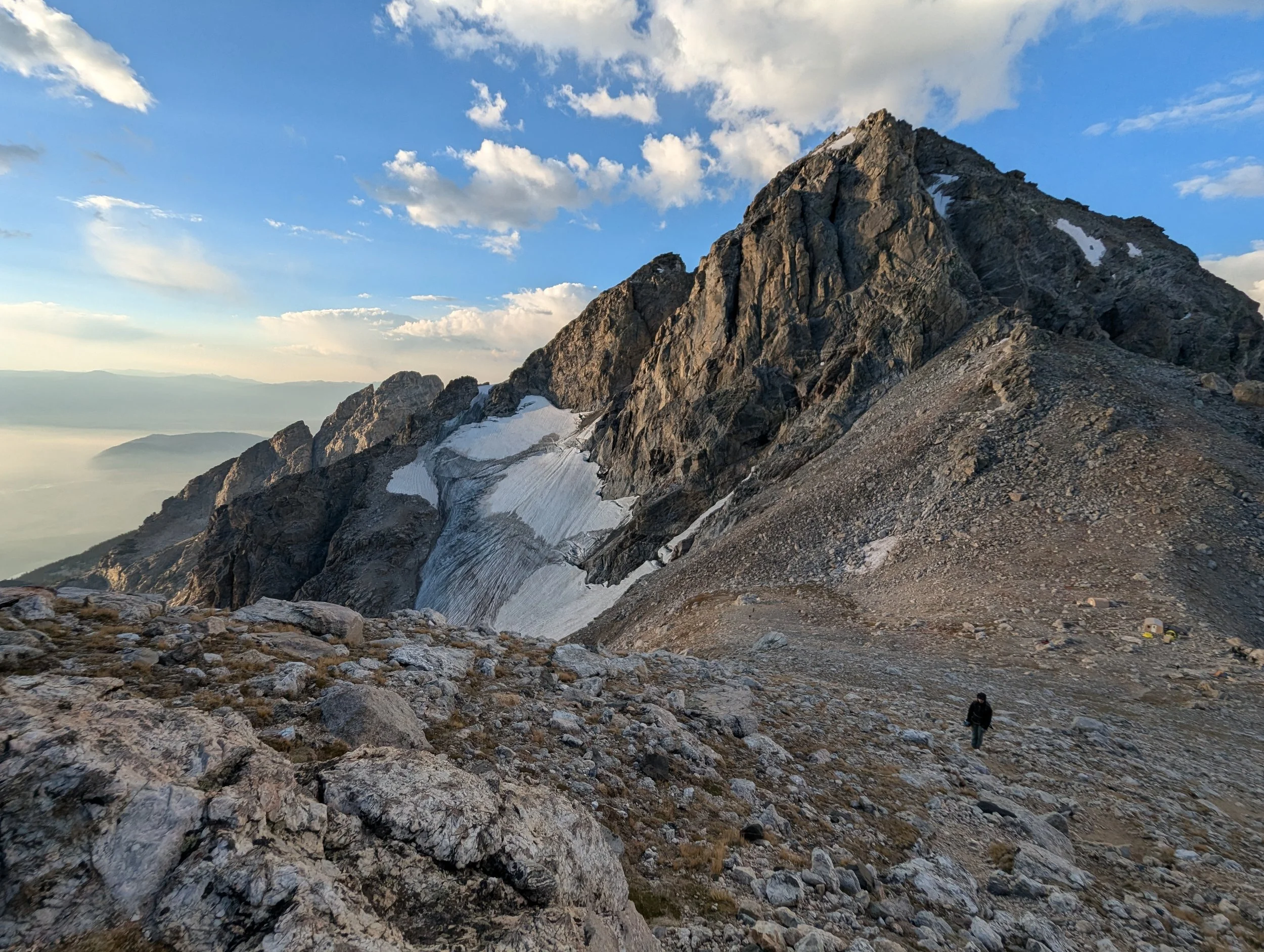 A person hiking on a rocky mountain trail with a large mountain peak in the background during daytime.