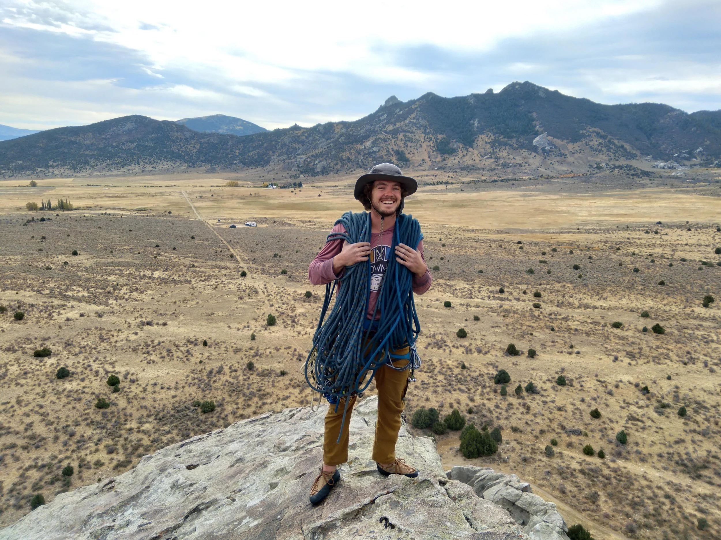 A man standing on a rocky peak holding climbing ropes with a vast open plain and mountains in the background, smiling at the camera.