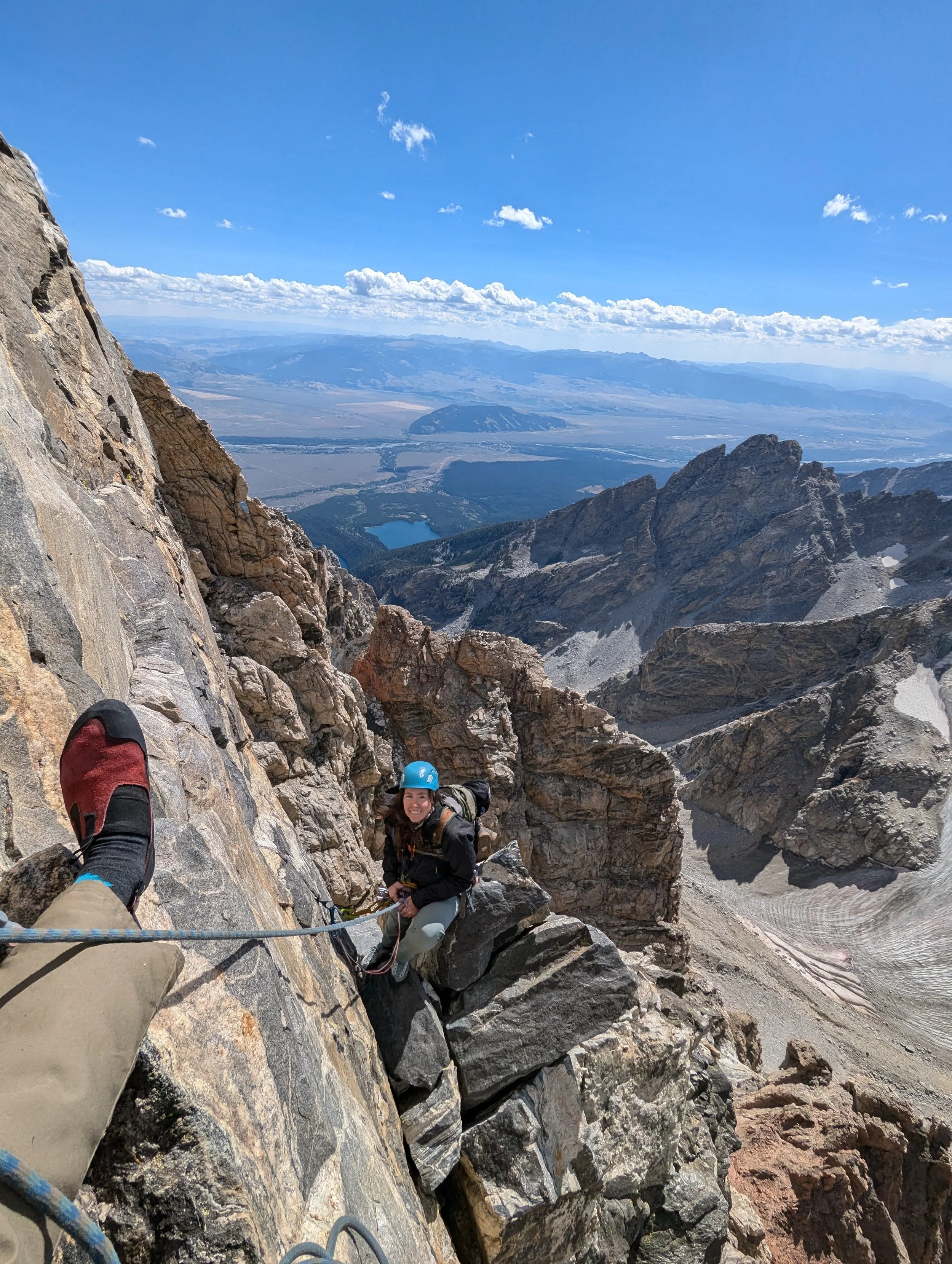 A person rock climbing on a steep mountain face with another person visible in the background, overlooking a vast mountainous landscape with clouds in the sky.