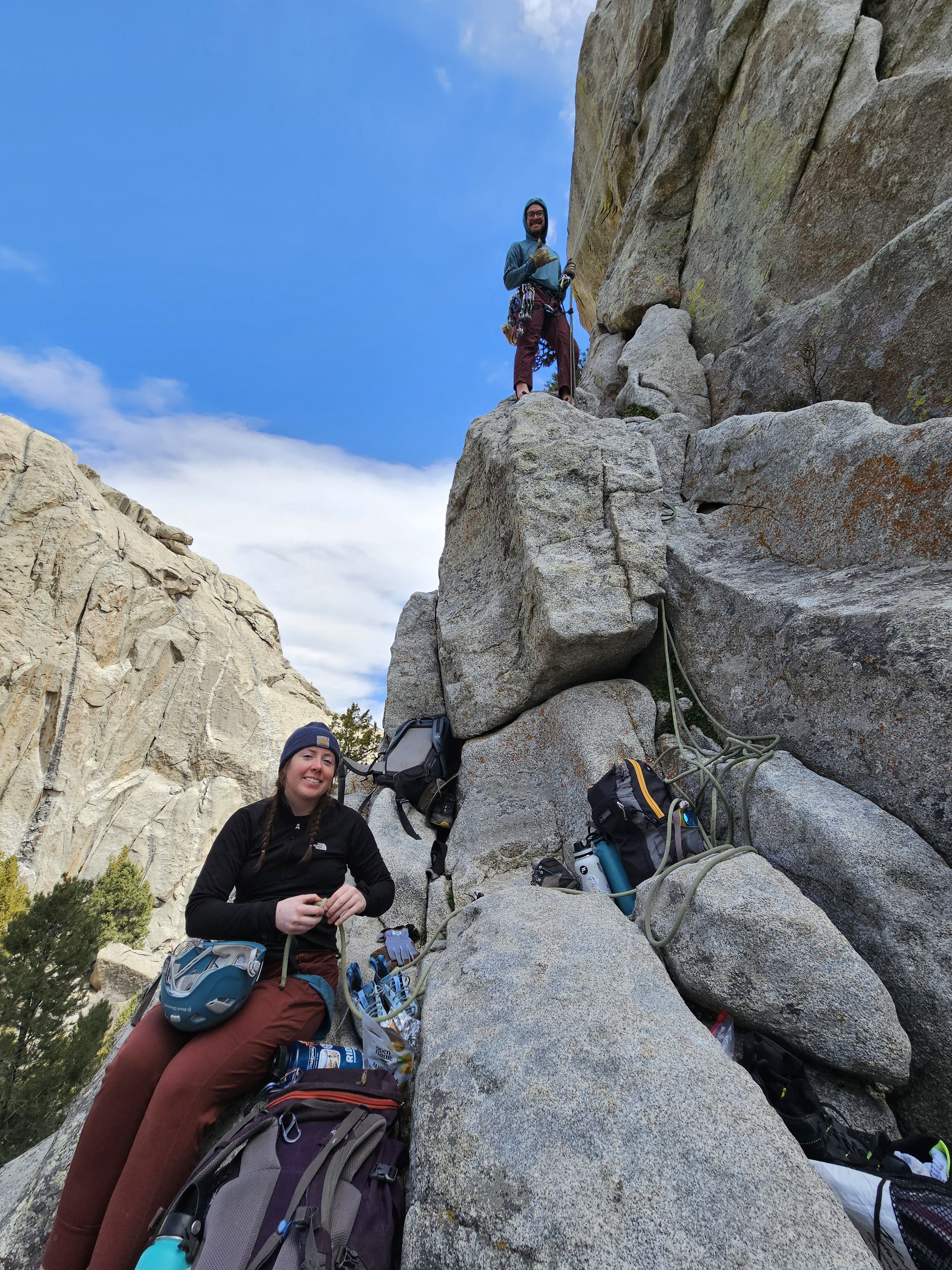 Two women rock climbing on a steep mountain with backpacks and climbing gear, one standing on a large boulder above, the other sitting on a rock at the base, surrounded by outdoor equipment and rugged terrain.