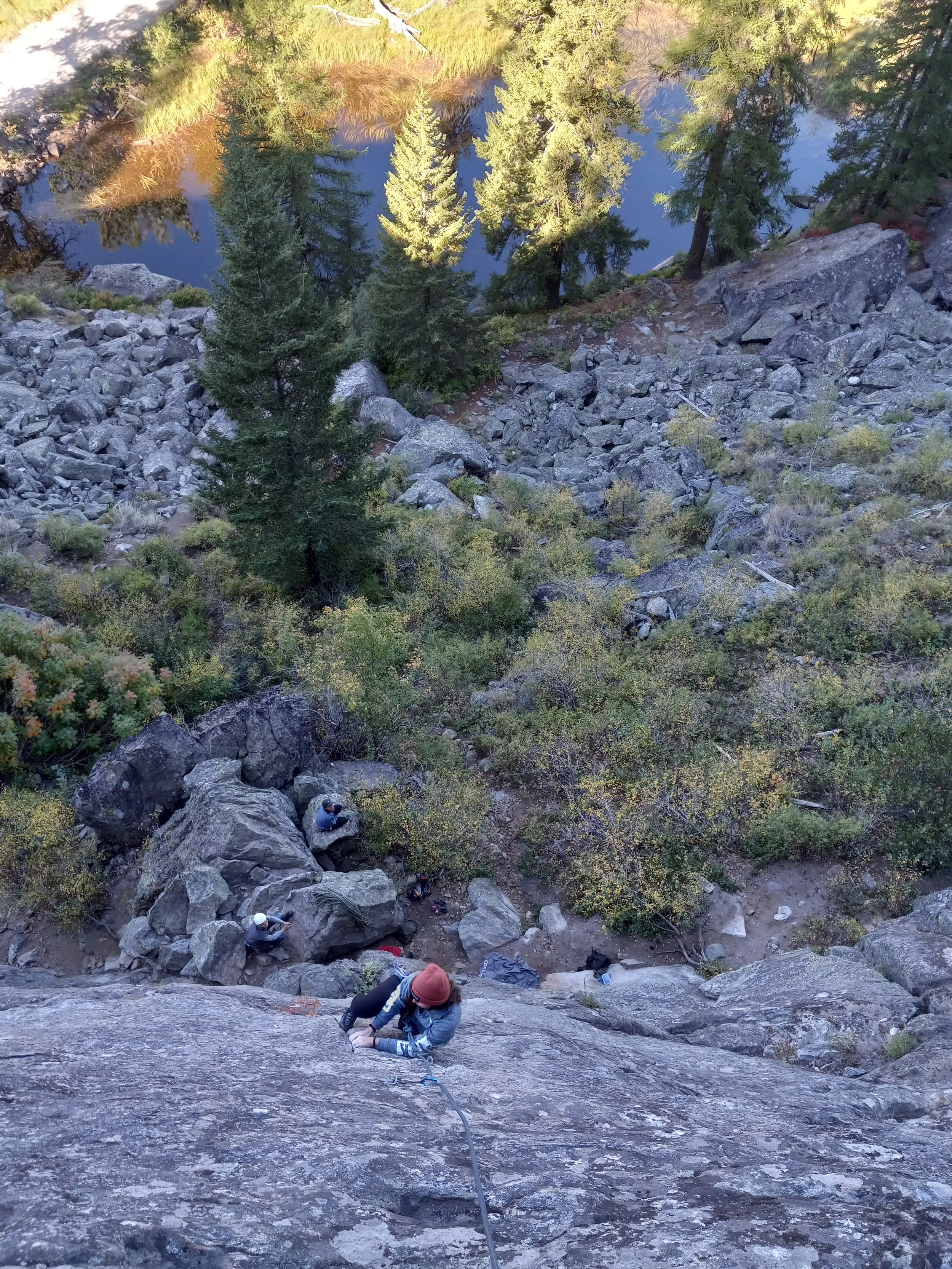 A person rock climbing on a steep granite wall with a safety harness, with two other climbers resting on rocks below, surrounded by trees and a body of water in the background.