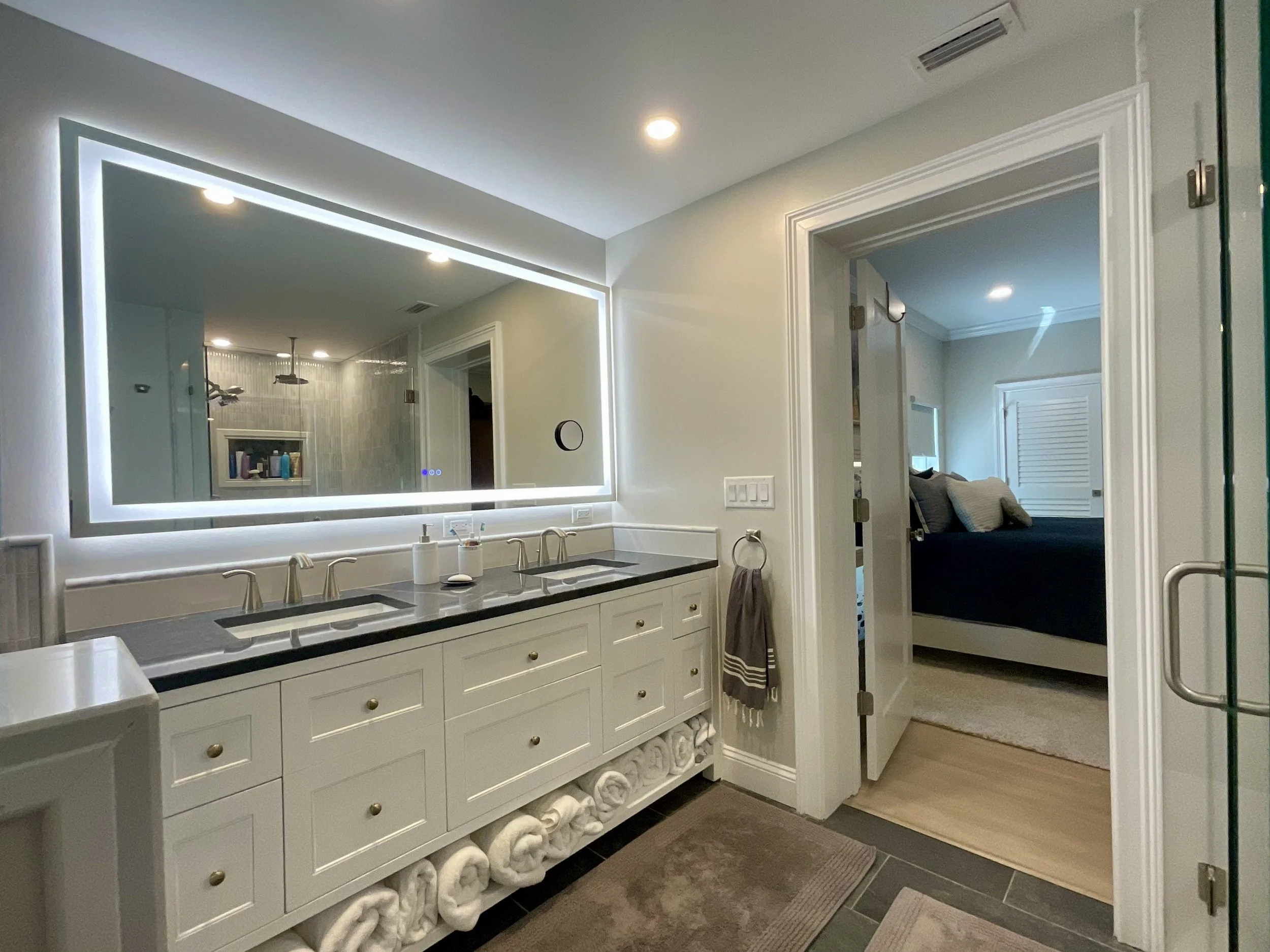Double-sink vanity with large illuminated mirror in master bathroom, view toward door leading to bedroom.
