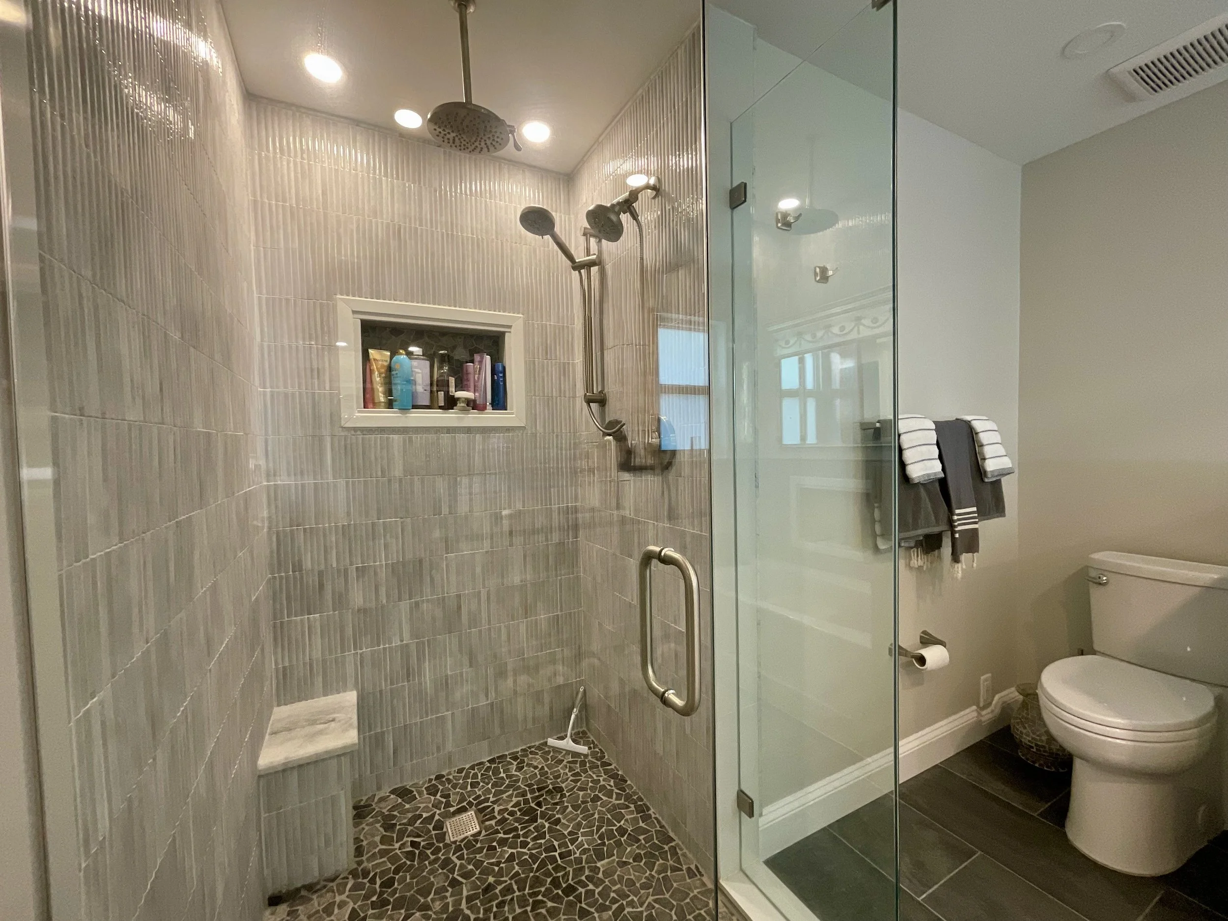 Corner shower with glass walls, floor-to-ceiling tile, rainshower, regular shower head, handheld spray, niche, and corner seat; toilet with privacy half wall.
